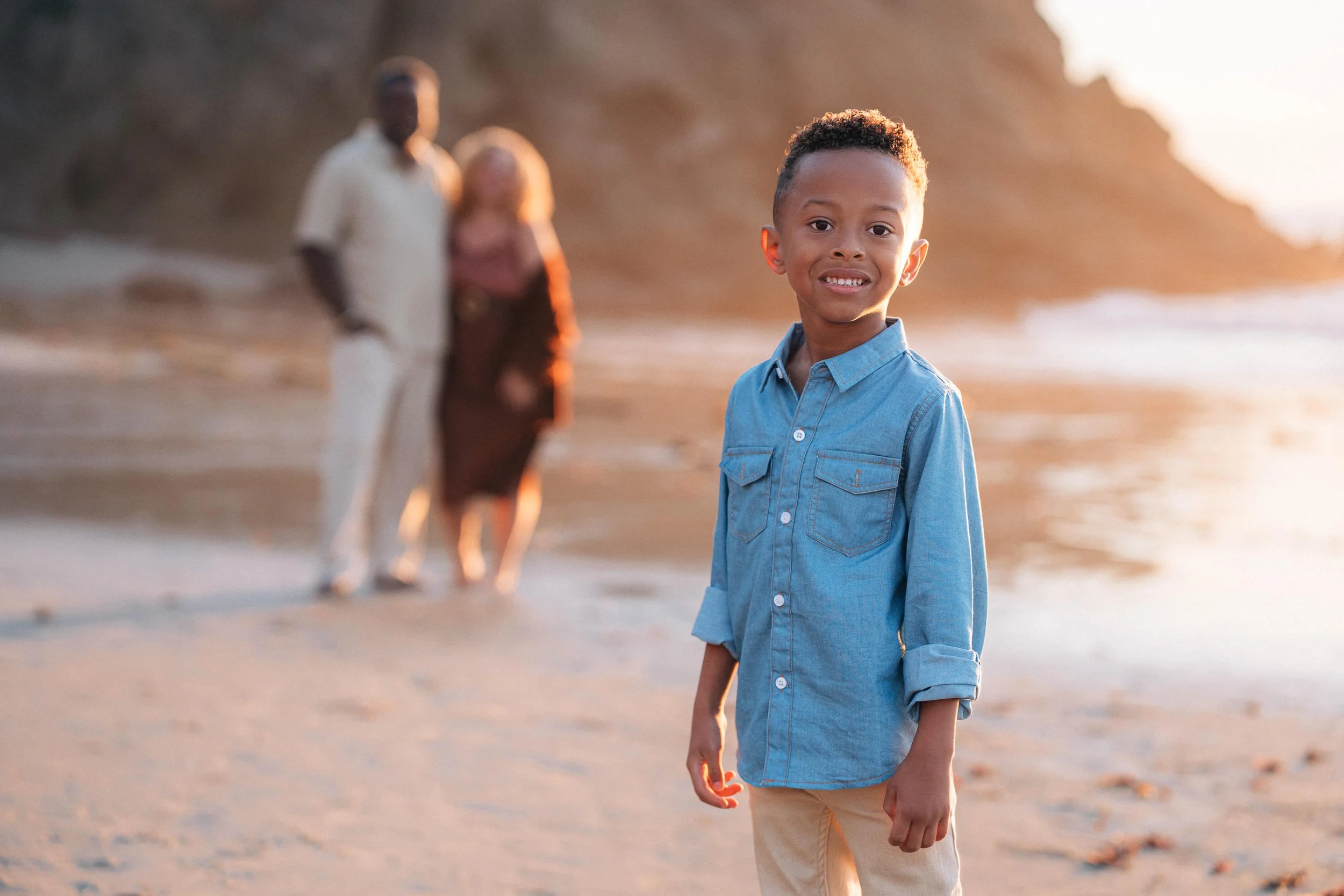 A young boy smiling at the camera on a beach during sunset, with two adults blurred in the background, walking along the shoreline.