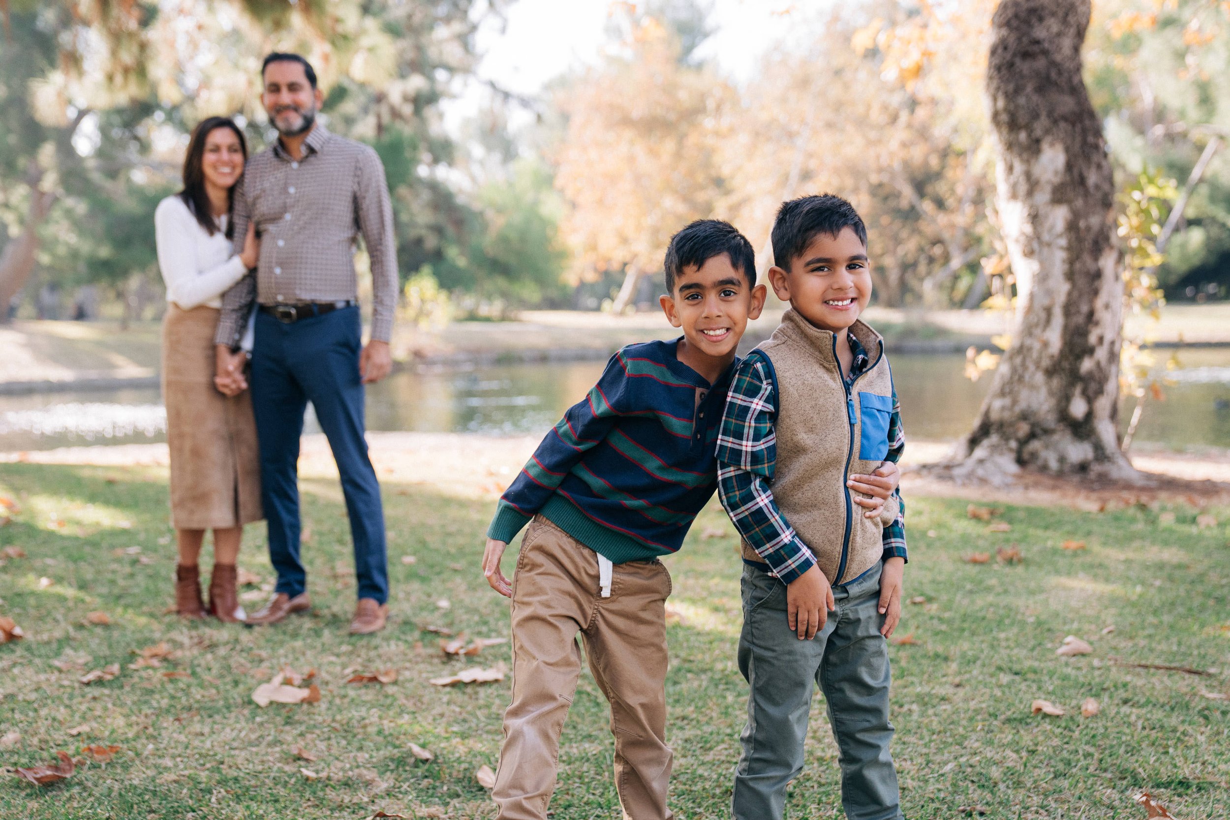 Two young boys smiling and standing close together in front of a tree and a body of water. In the background, a woman and a man are holding hands and smiling, standing on a grassy area with fallen leaves and trees with autumn foliage.