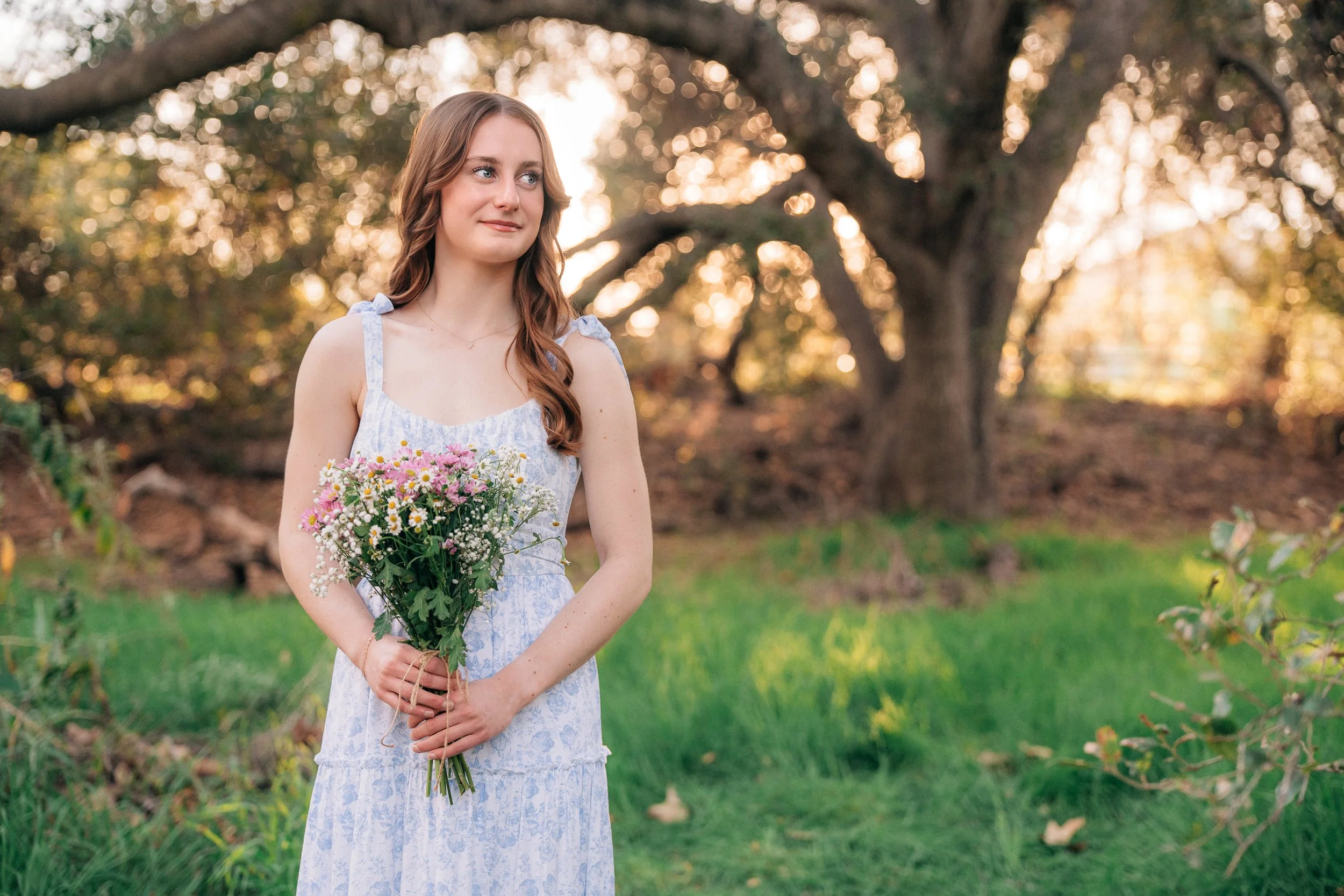 A young woman with long brown hair in a white and blue dress holding a bouquet of flowers, standing outdoors in a grassy area with trees in the background and sunlight filtering through the leaves.