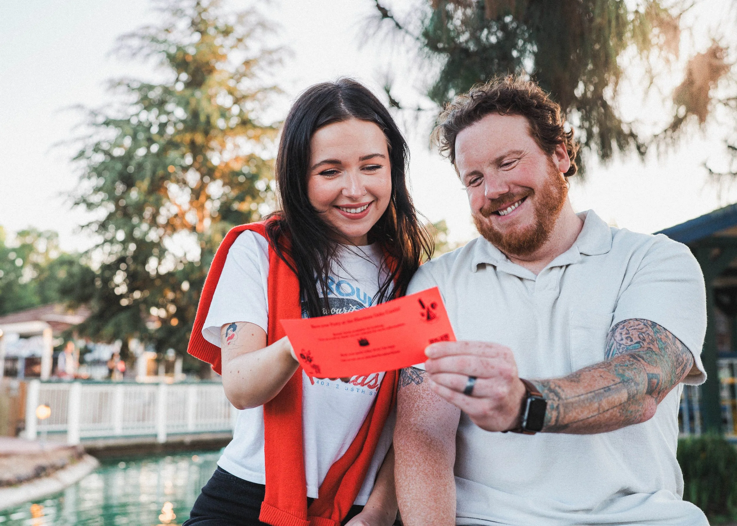A smiling woman with black hair and a man with a beard, both casually dressed, looking at a bright red ticket or flyer together outdoors.