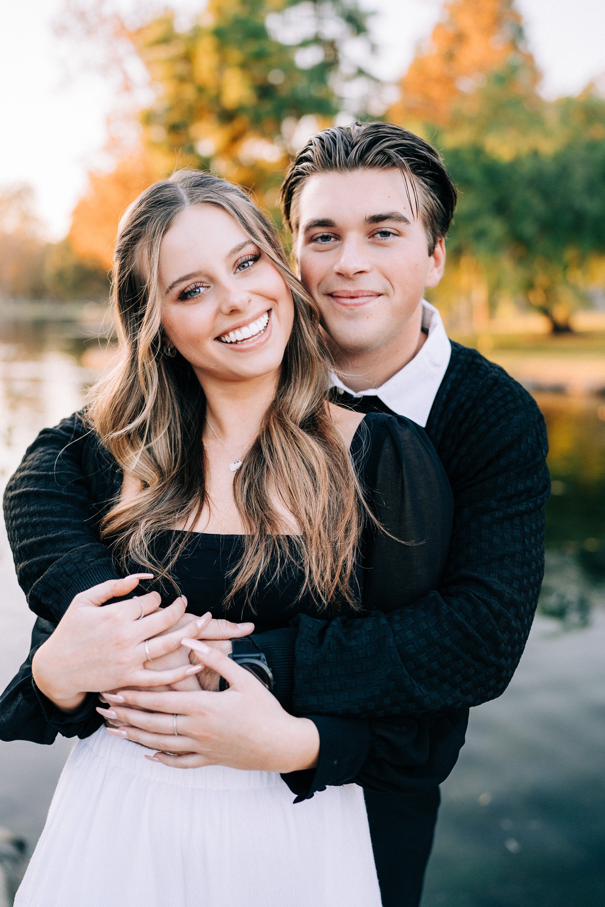 A happy couple embracing outdoors during autumn, with trees showing fall foliage in the background.