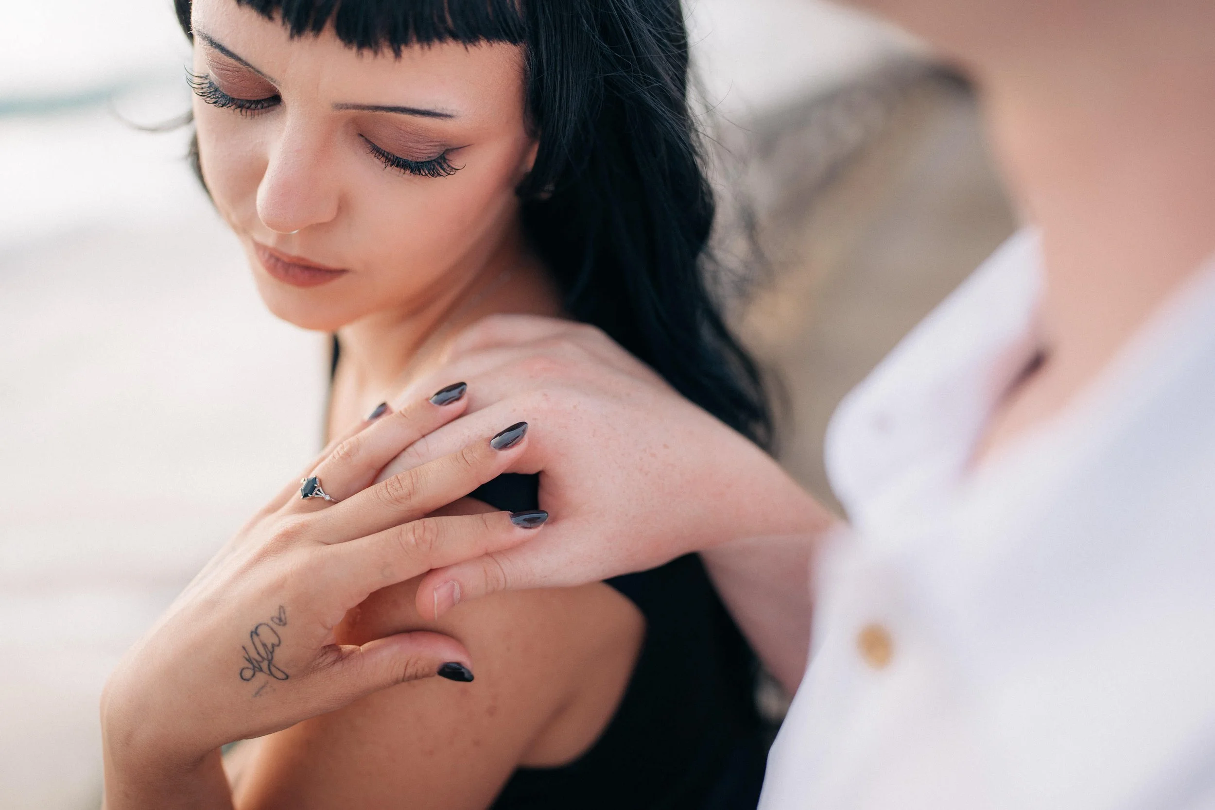 A woman with dark hair and makeup touching her shoulder, showing a tattoo and a ring, with a blurred arm and part of a white shirt visible.