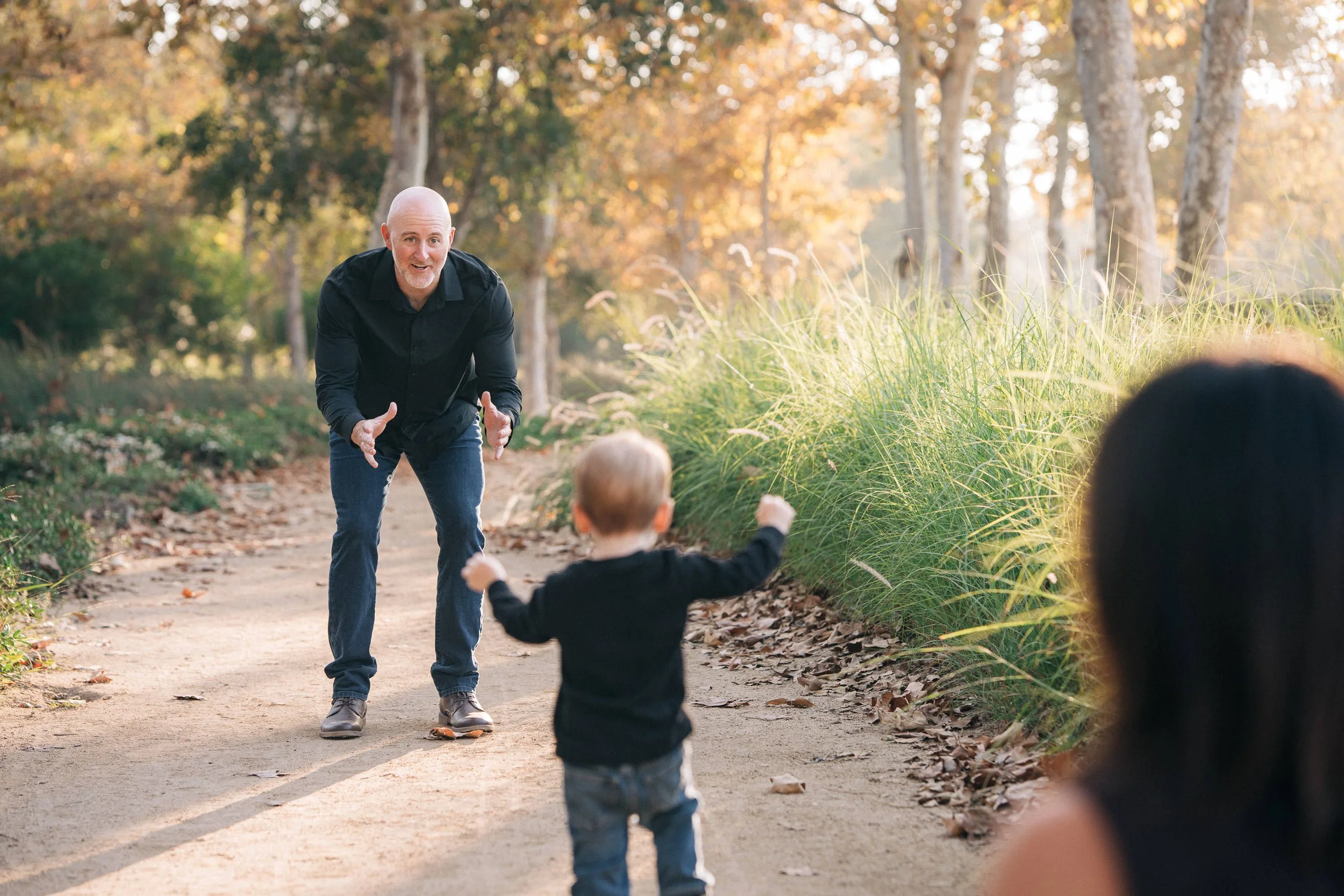 A man with a bald head and beard wearing a black shirt and jeans is smiling and gesturing towards a young boy in a black shirt and jeans who is running on a dirt path in a wooded park during fall. A woman with dark hair, partially visible, is sitting