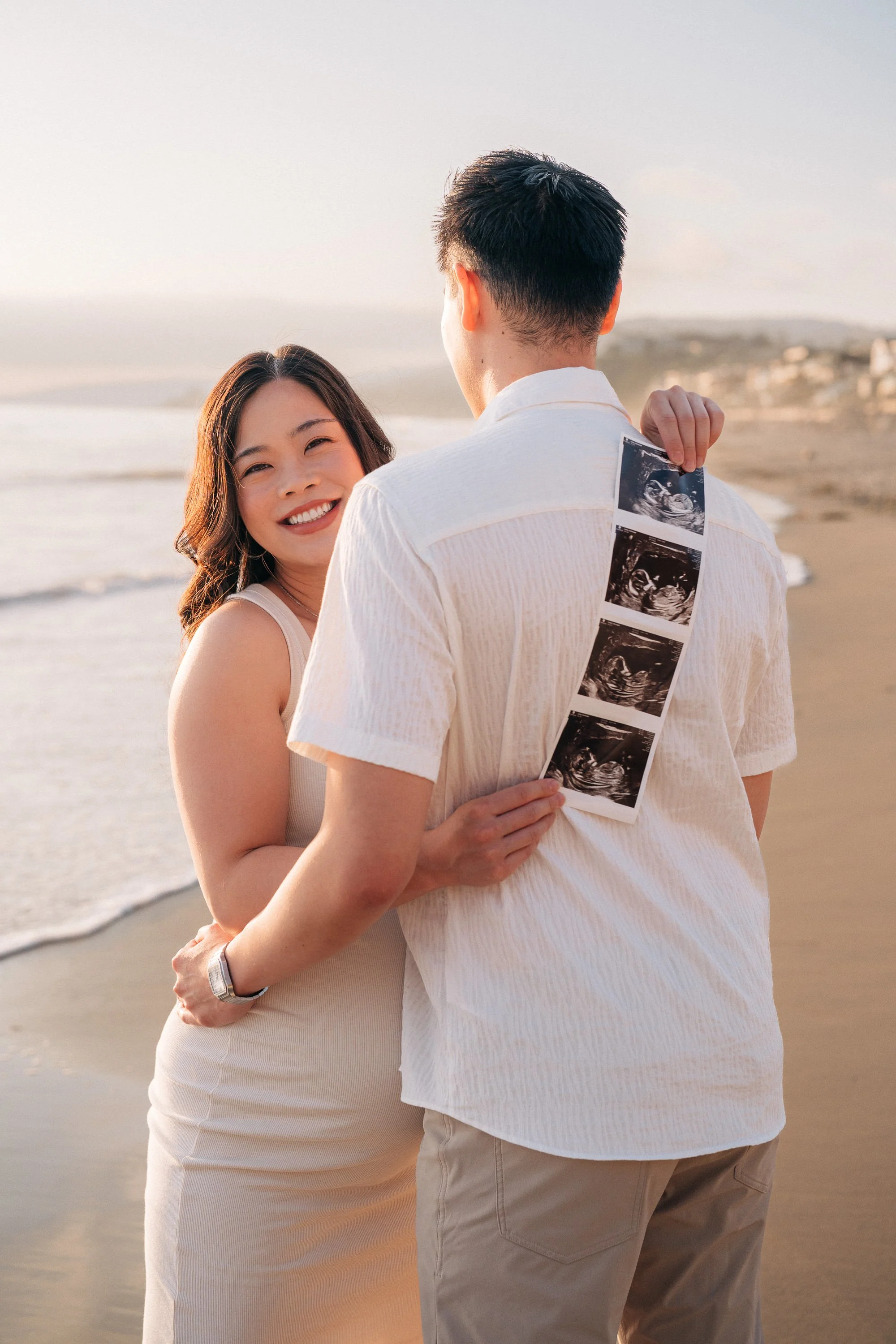 A couple on the beach, with the woman smiling while holding the man, who has ultrasound photos hanging from his back.