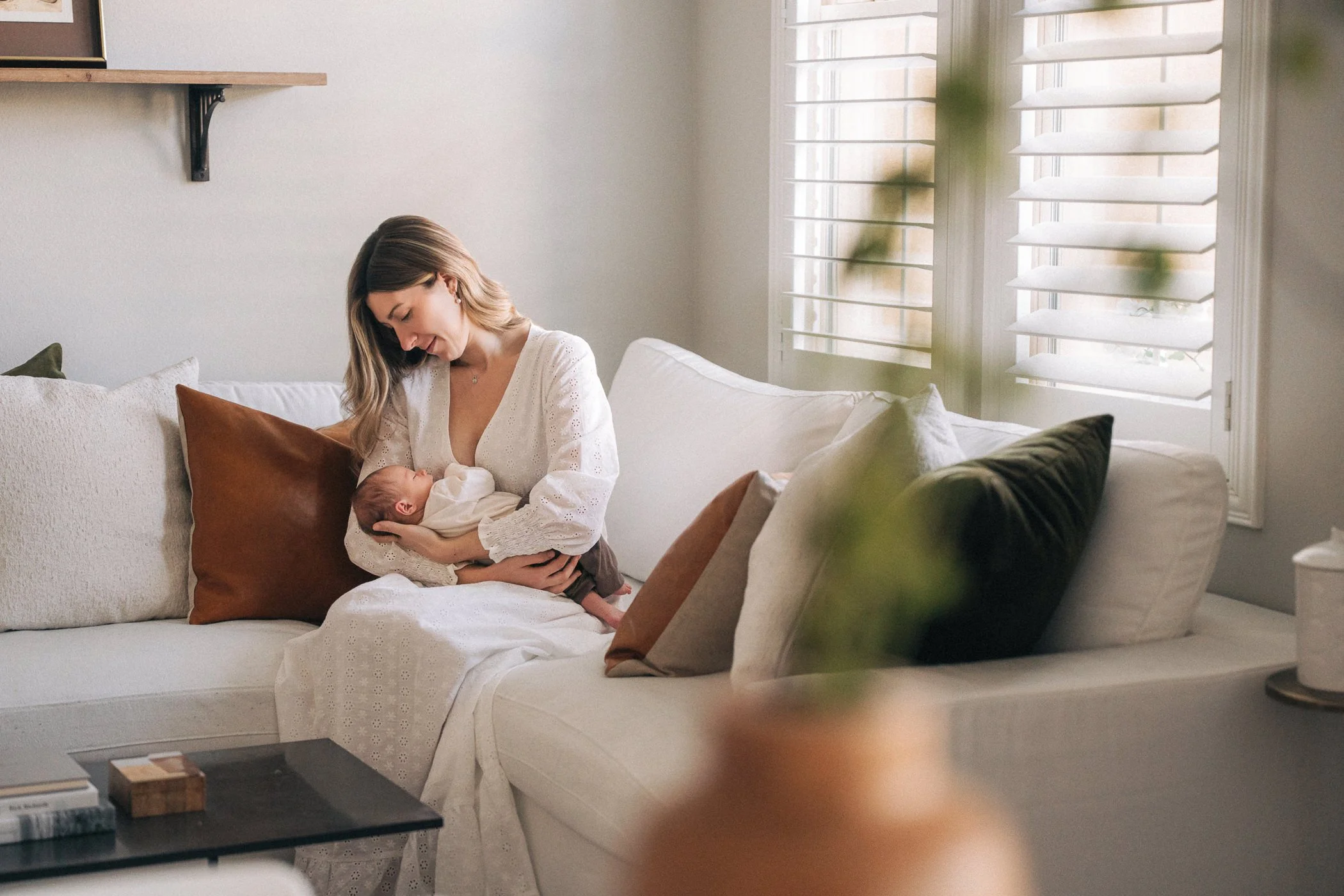 A woman with shoulder-length hair, wearing a white dress, sitting on a white couch, holding a newborn baby in her arms, in a bright living room with large windows, blinds, and decorative pillows.