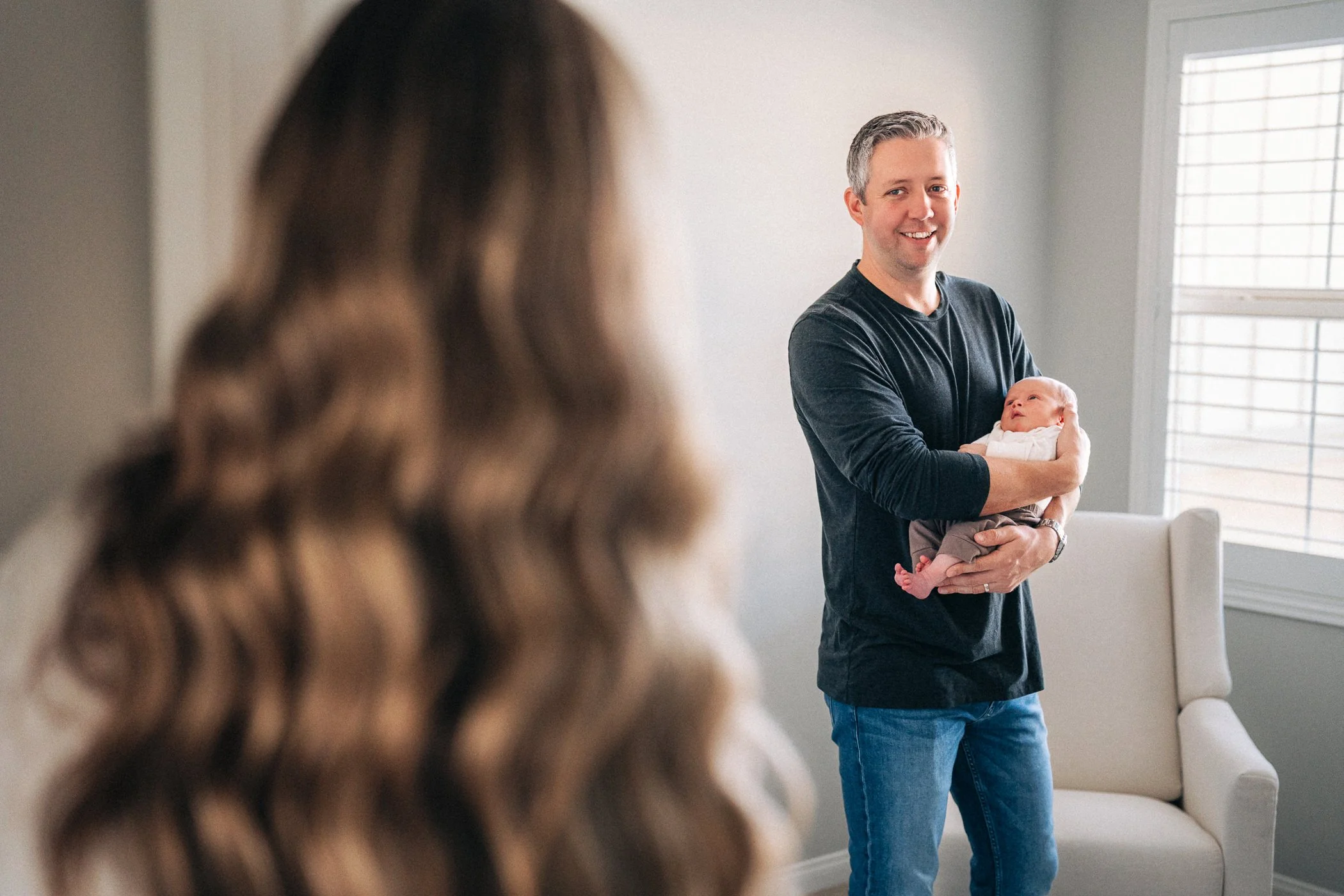 A man holding a newborn baby, smiling indoors with a woman in the foreground out of focus.