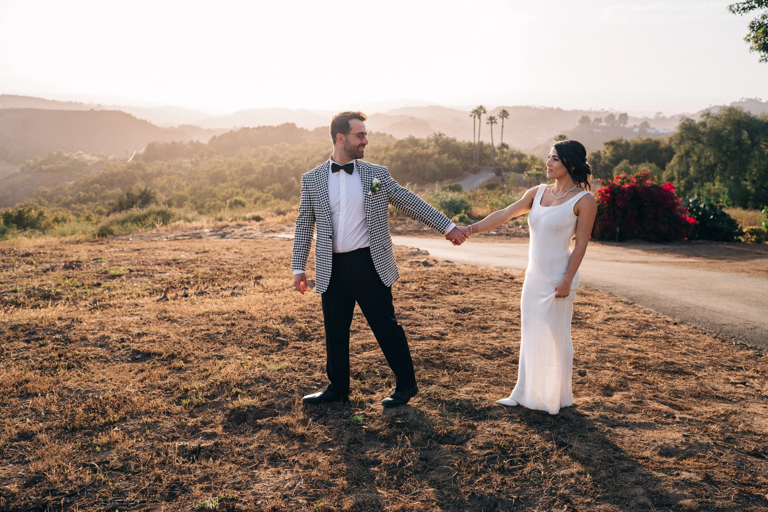 A newlywed couple holding hands and smiling outdoors at sunset, with a scenic hilly background, some greenery, and flowering bushes.