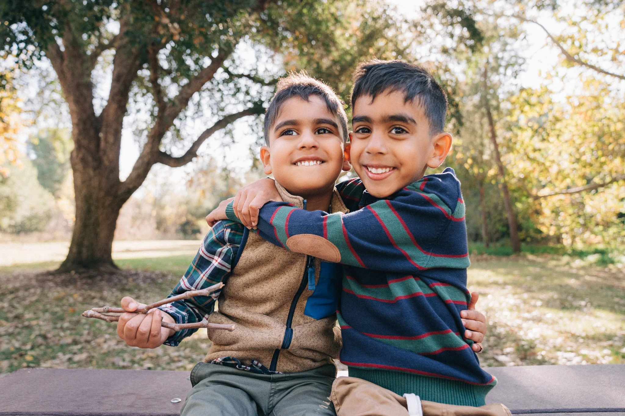 Two young boys with short dark hair and brown skin hugging and smiling outdoors during autumn, with one boy holding a stick, standing in front of trees with yellow and orange leaves.