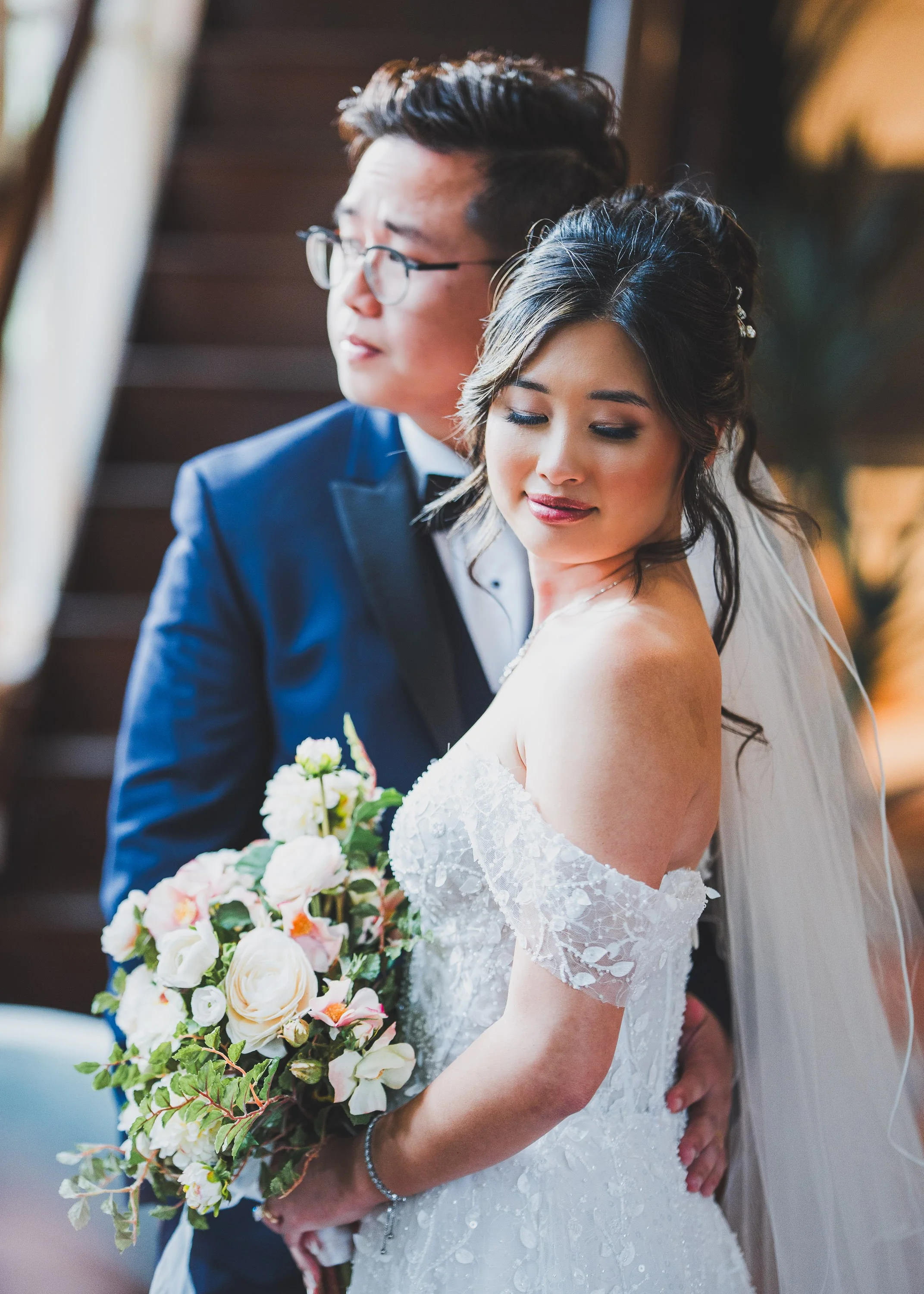 A bride in a lace wedding dress and veil holding a bouquet of white and pink flowers, standing closely with a groom in a navy suit and glasses, both with their eyes closed in an intimate moment.