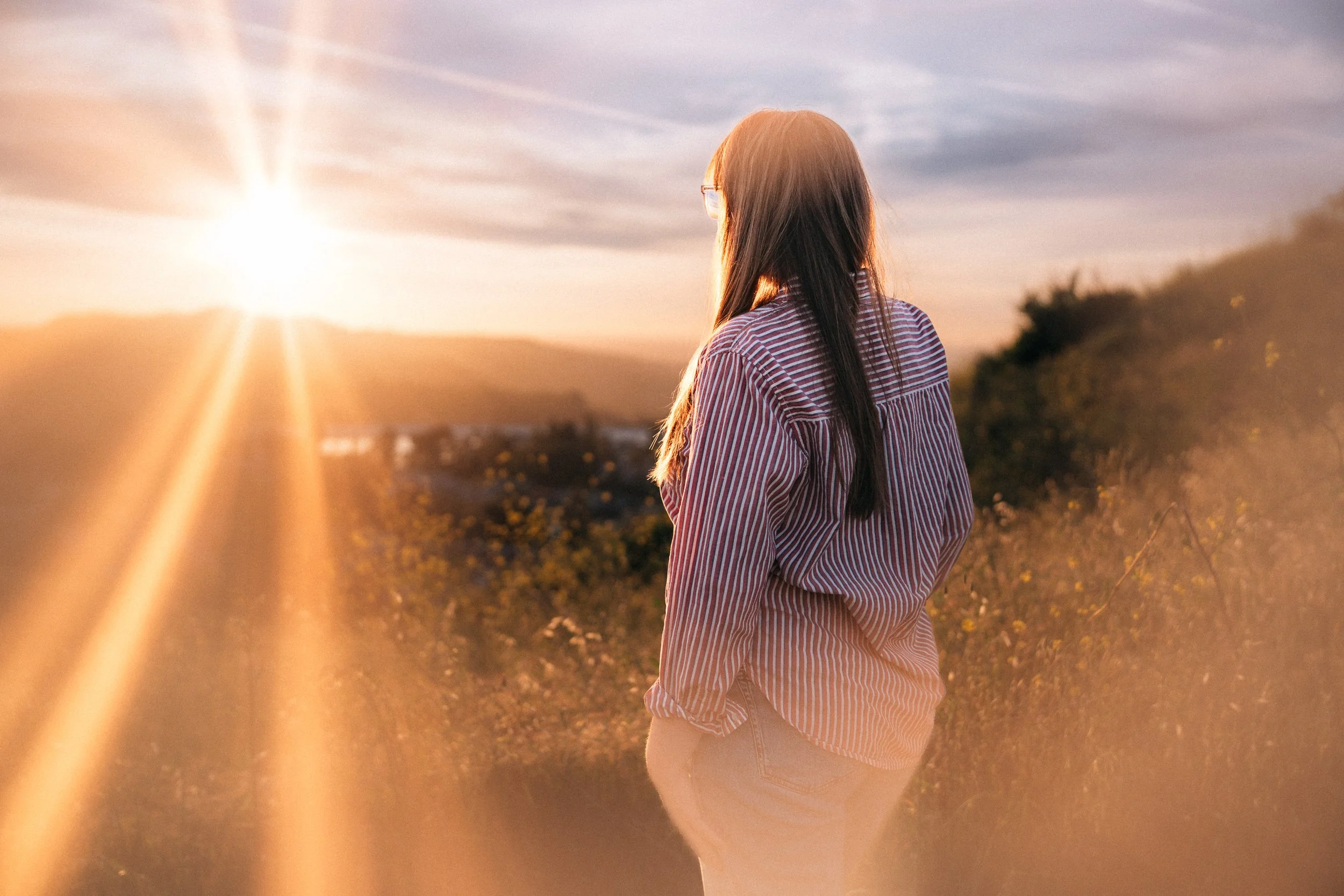 Woman with long dark hair wearing glasses, striped shirt, and jeans, standing in a field at sunset, gazing towards the horizon.