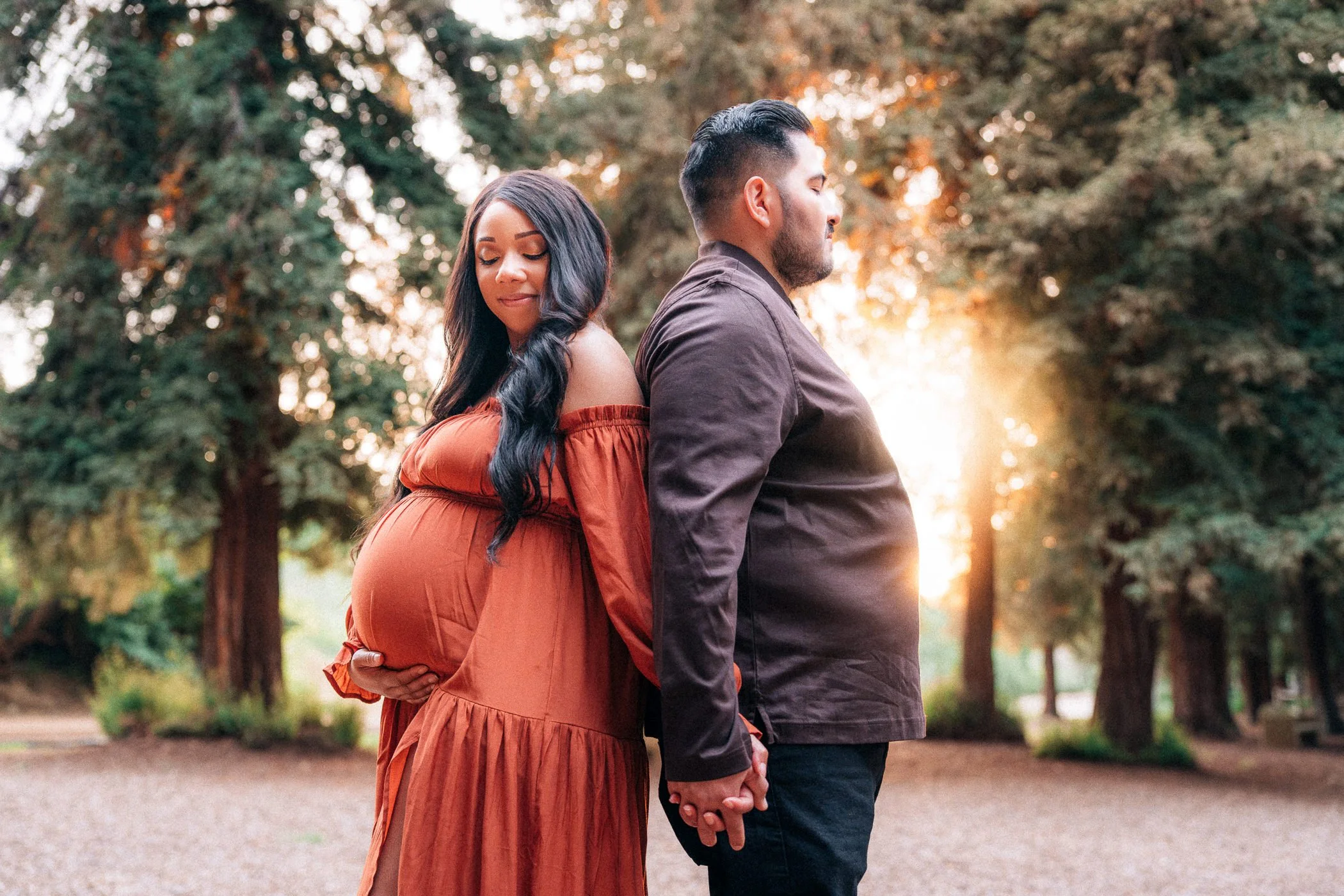 A pregnant woman and a man hold hands with their backs to each other in a park at sunset.