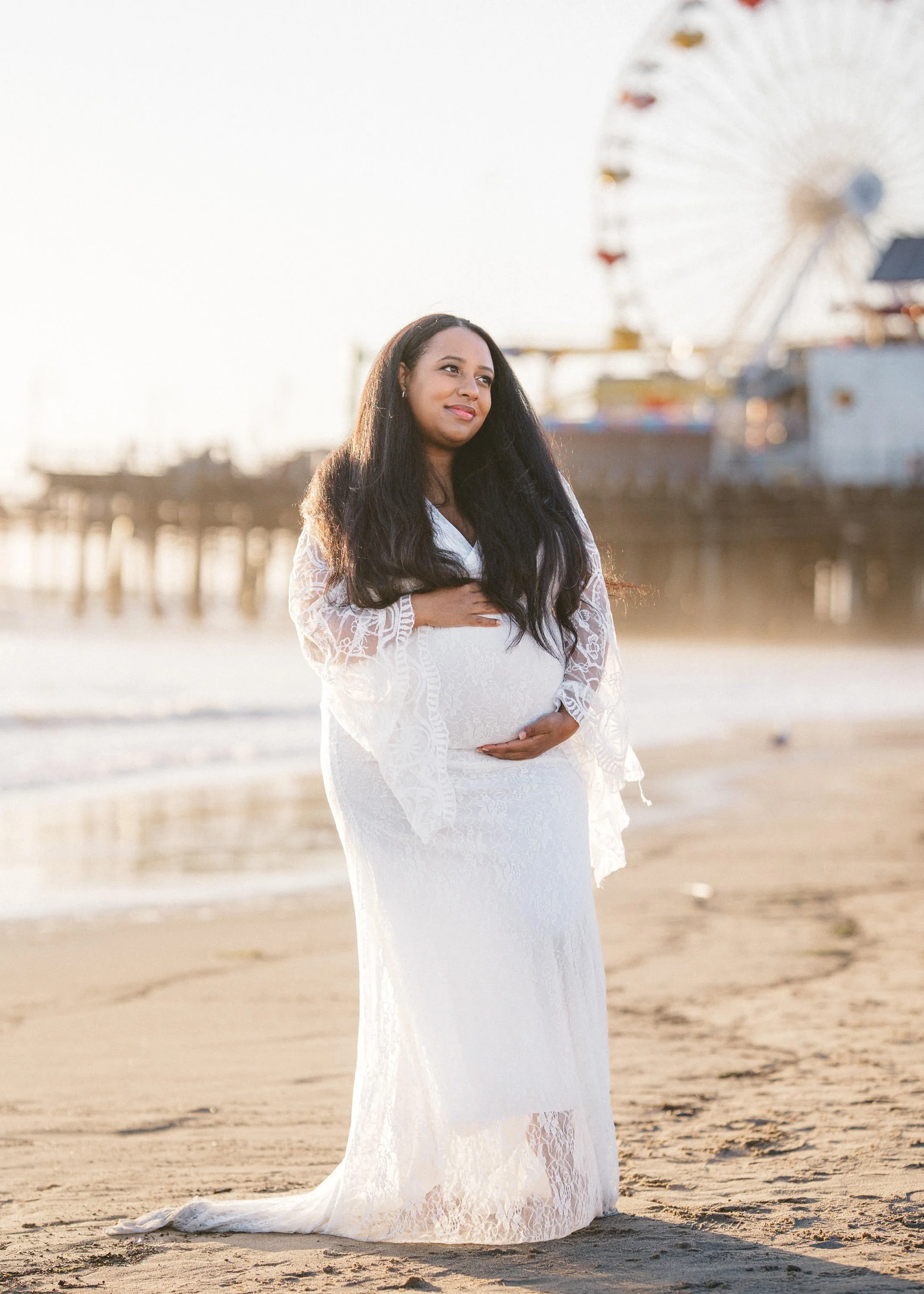A pregnant woman in a white lace dress standing on a beach, with a pier and Ferris wheel in the background during sunset.