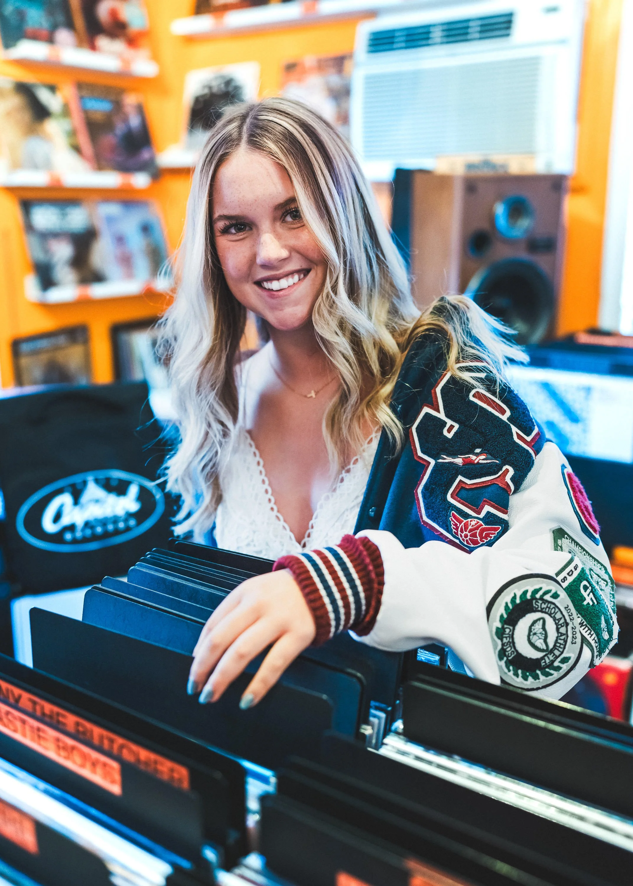 A young woman with long blonde hair smiling at the camera, sitting at a record store or music shop.