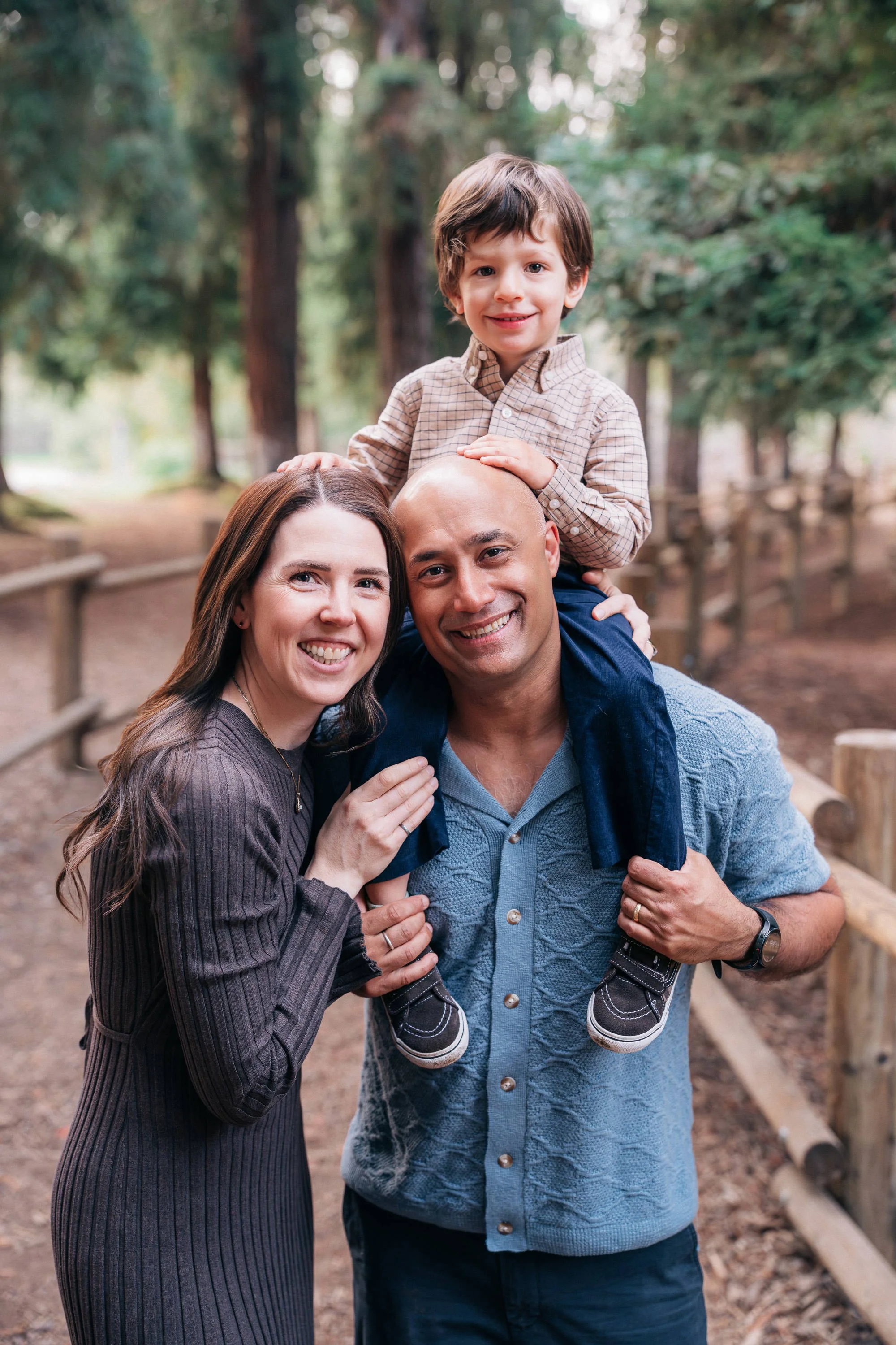 Three people, a woman, a man, and a young boy, smiling and enjoying a walk in a wooded park. The boy is sitting on the man's shoulders, and the woman is standing beside them, holding the boy's foot.