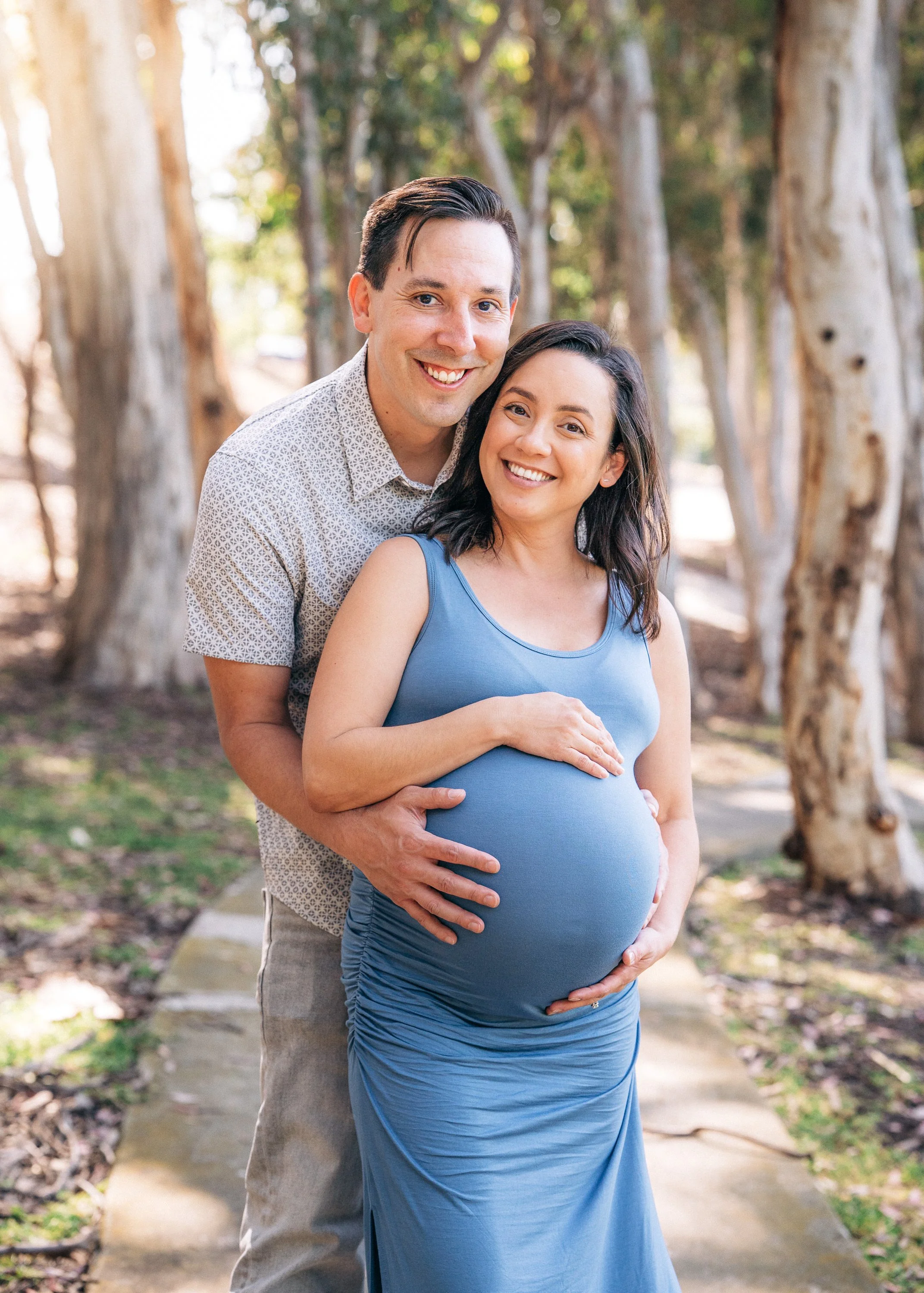 Smiling pregnant woman in a blue dress with her partner outdoors among trees, with partner's hands resting on her belly.
