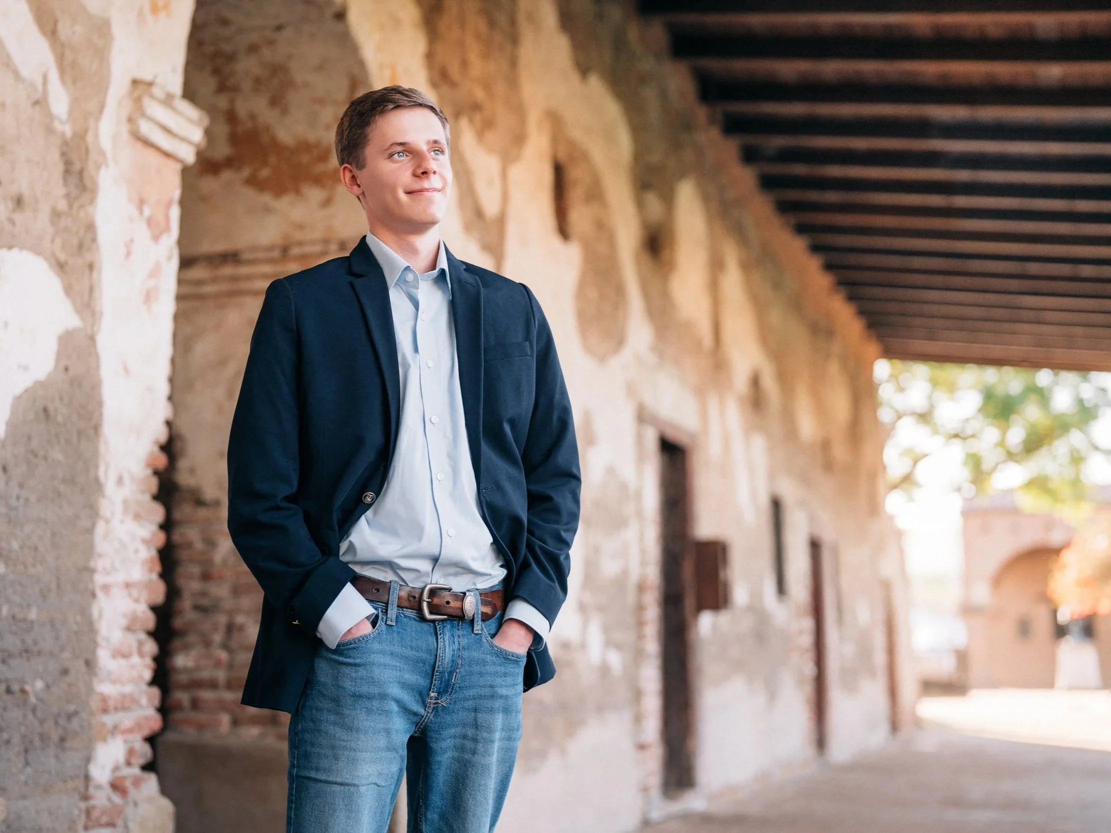 A young man in a blue blazer and jeans standing outdoors next to a brick wall, looking up and smiling.