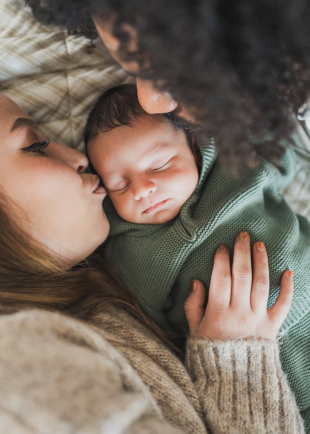 Mom kissing newborn's cheek as dad leans in close, baby in green knit sleeper, Nathan and Ana in-home session San Diego