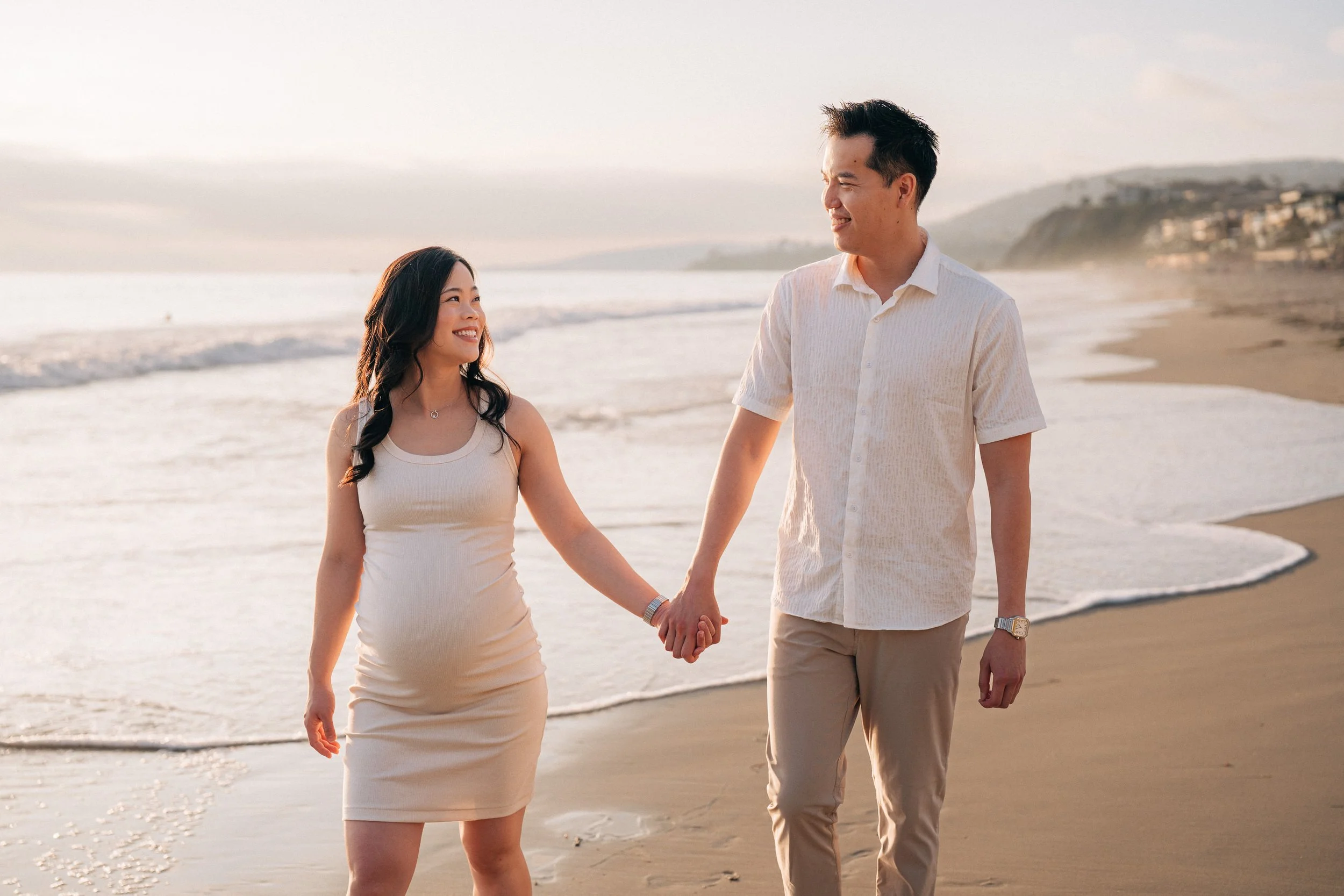 A couple holding hands and walking on the beach during sunset, smiling at each other.