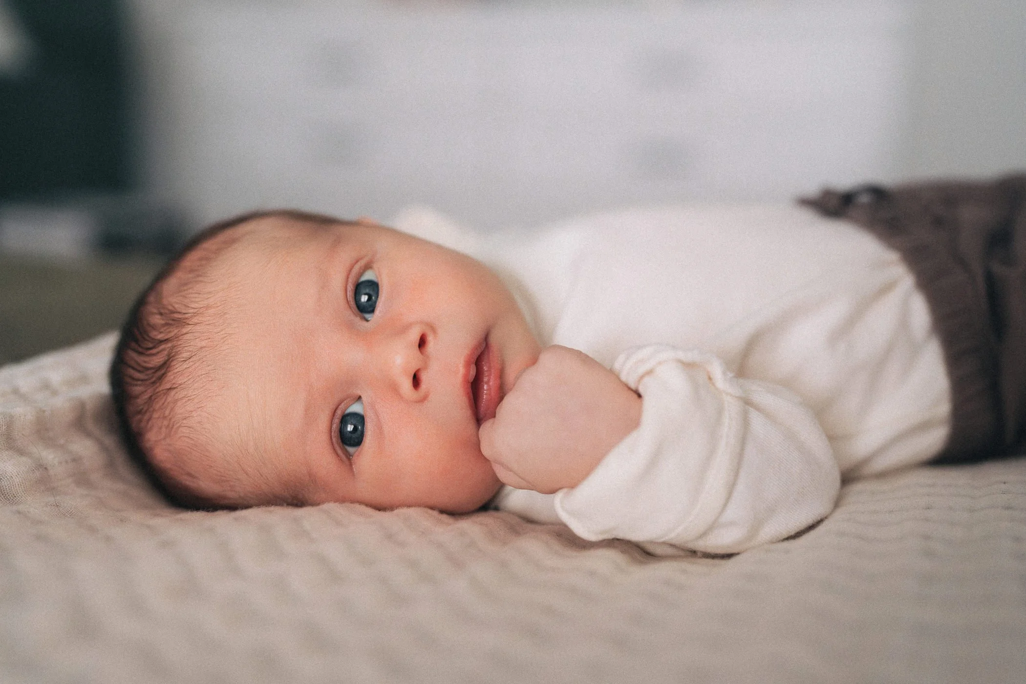 Close-up of a baby lying on a soft surface, looking at the camera with blue eyes, wearing a white long-sleeve shirt, in a relaxed pose with hand near mouth.