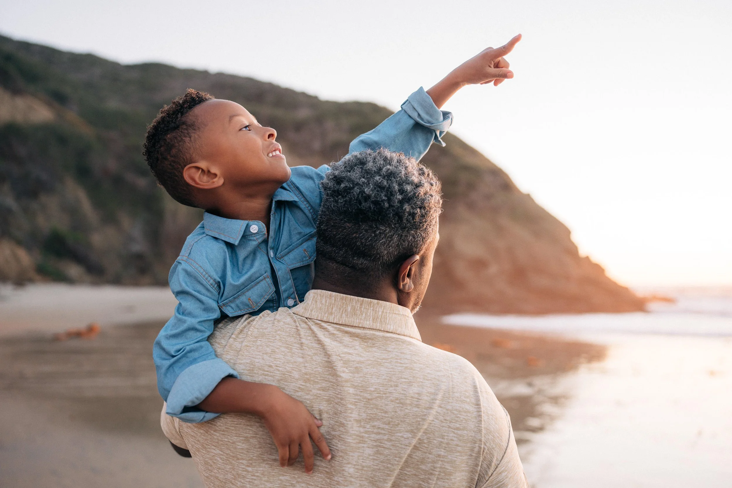 A young boy sitting on an adult's shoulder on a beach, pointing towards the ocean with a cliff in the background during sunset.