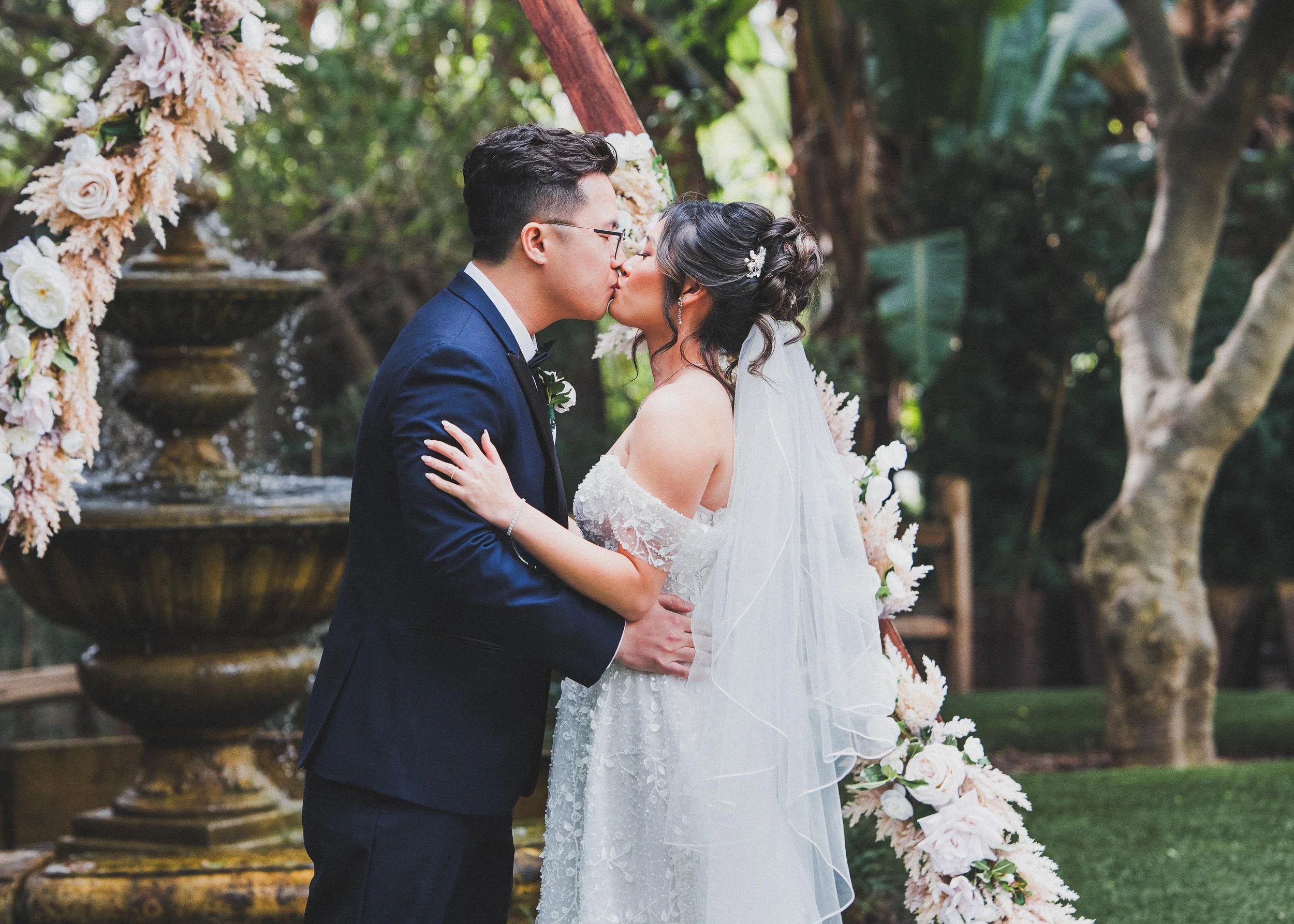 A wedding couple sharing a kiss outdoors, with a floral arch and water fountain in the background.