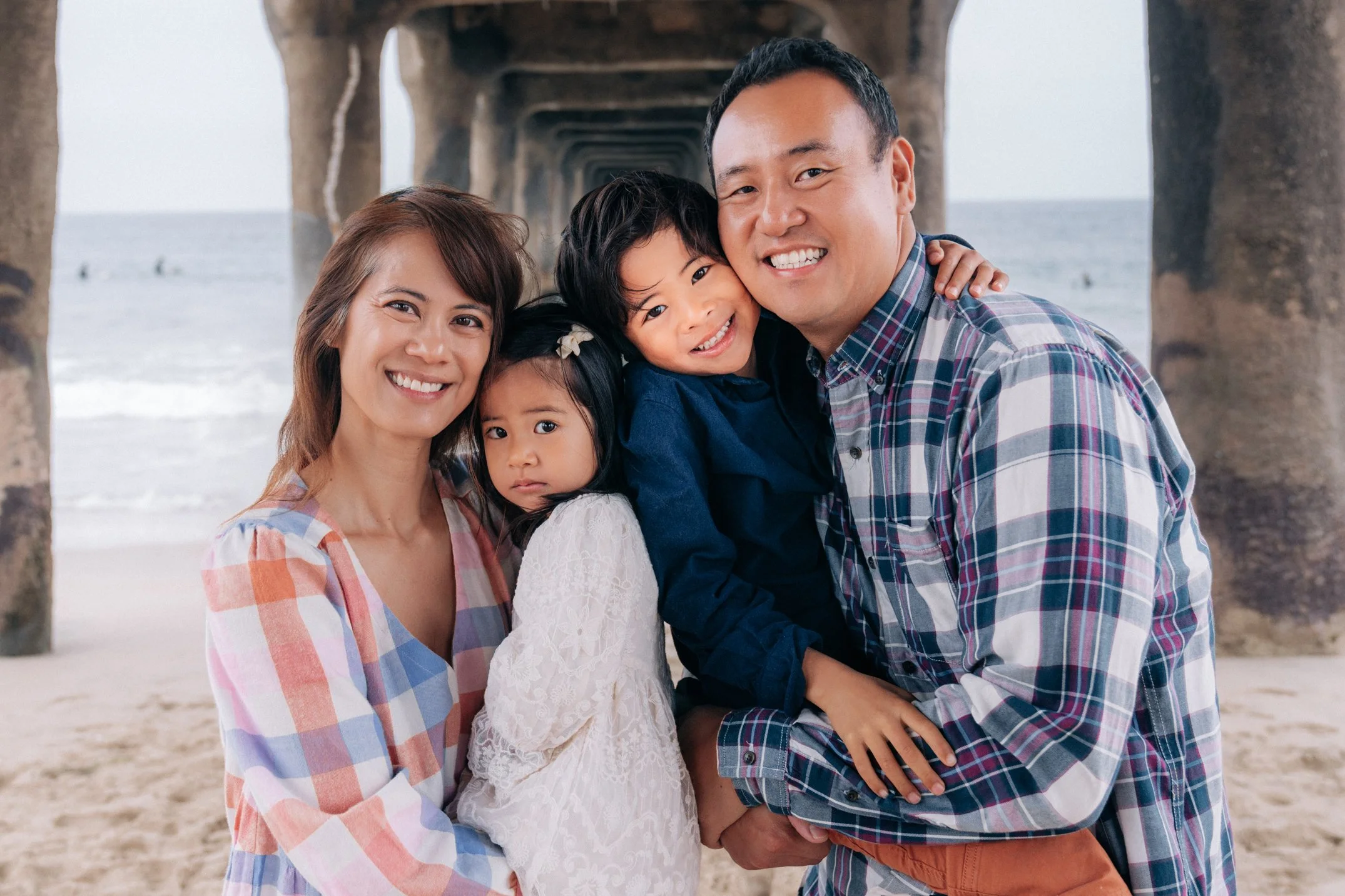 A happy family of five posing together on a beach pier, smiling at the camera with ocean in the background.