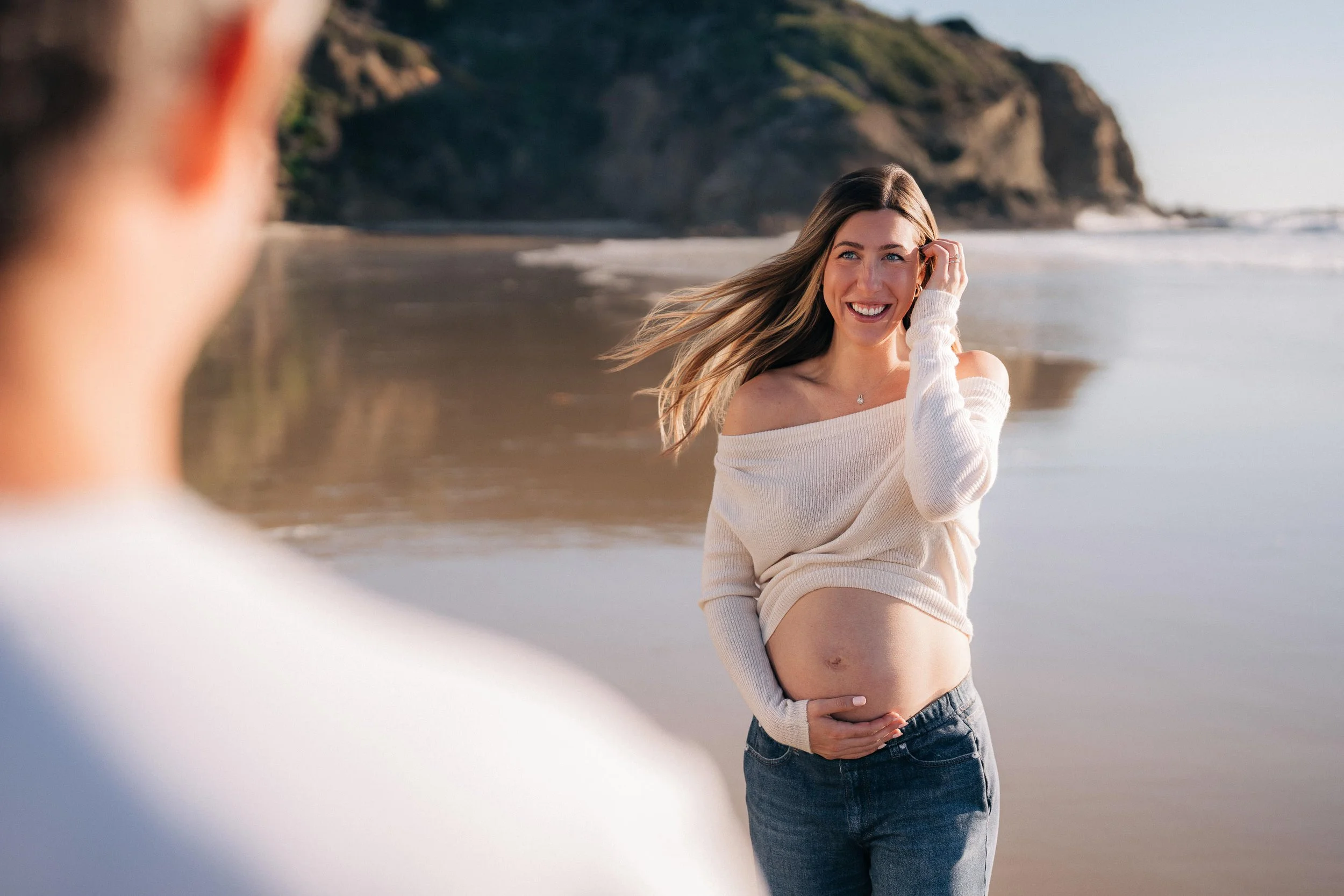 A pregnant woman smiling on a beach, touching her stomach, with wind in her hair and a man in the foreground, out of focus.