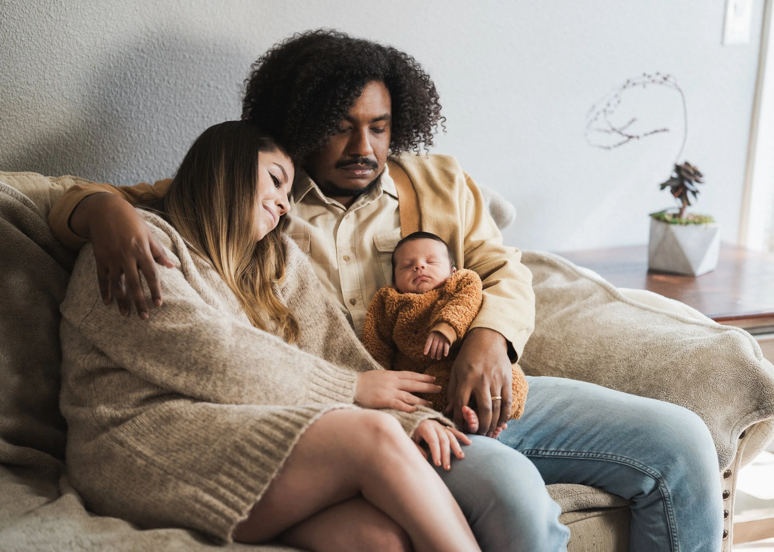 A family of three sitting on a beige sofa. The woman and man are holding a sleeping baby. The woman has long hair and is wearing a beige sweater; the man has curly hair and is wearing a light-colored shirt and beige cardigan. They look peaceful and c