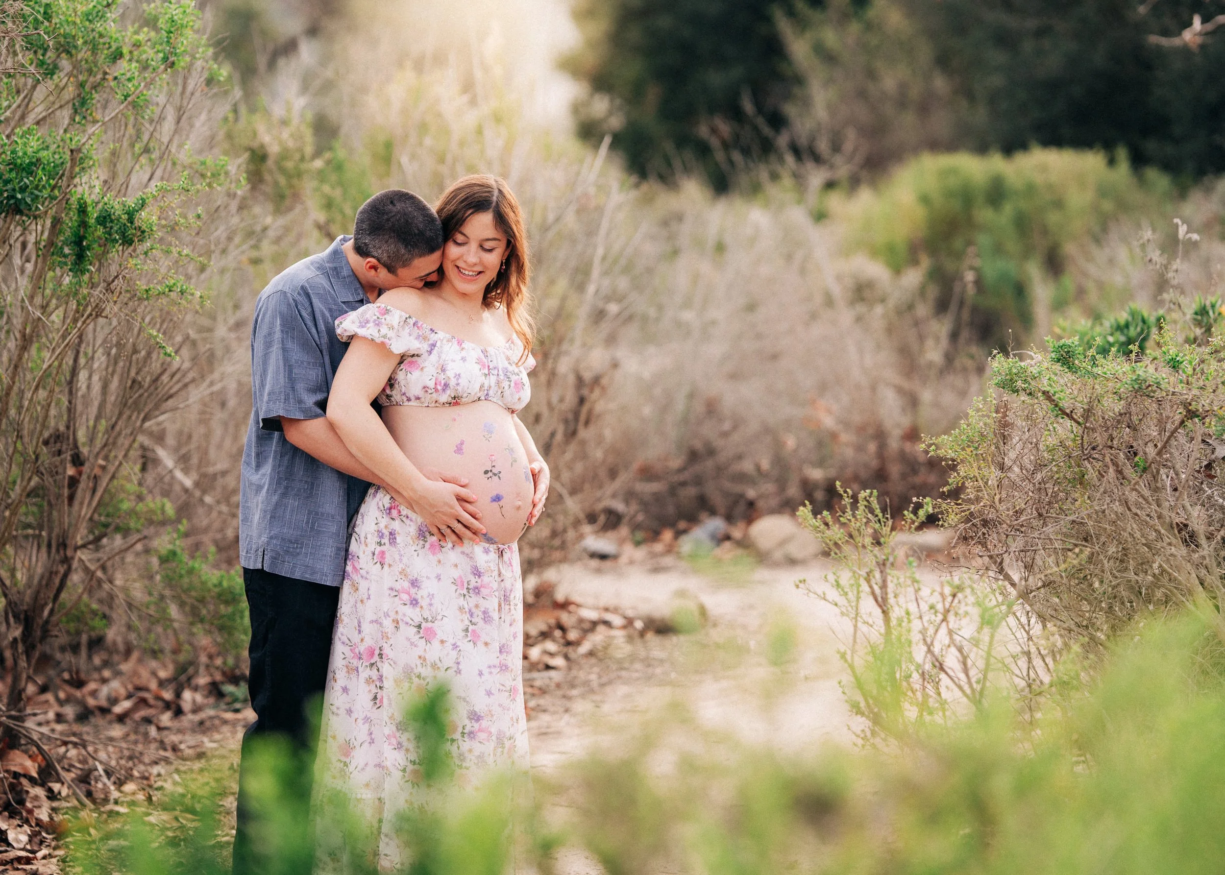 A pregnant woman in a floral dress is being embraced by a man in a blue shirt, both standing outdoors among bushes and trees. The man is kissing her cheek, and they are both smiling, celebrating her pregnancy.