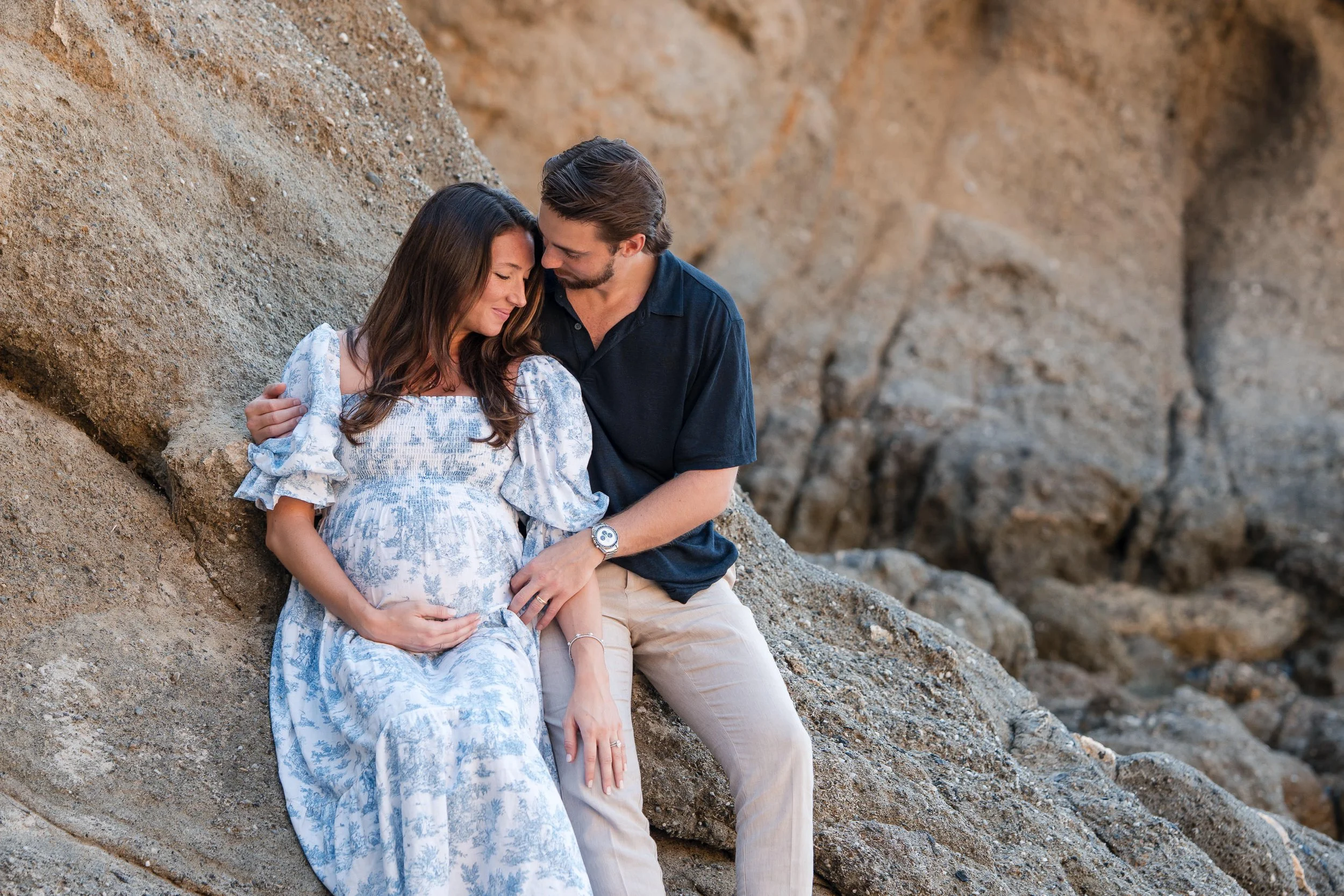 A couple sitting on rocks at the beach, woman is pregnant, they are holding hands and looking at each other lovingly.