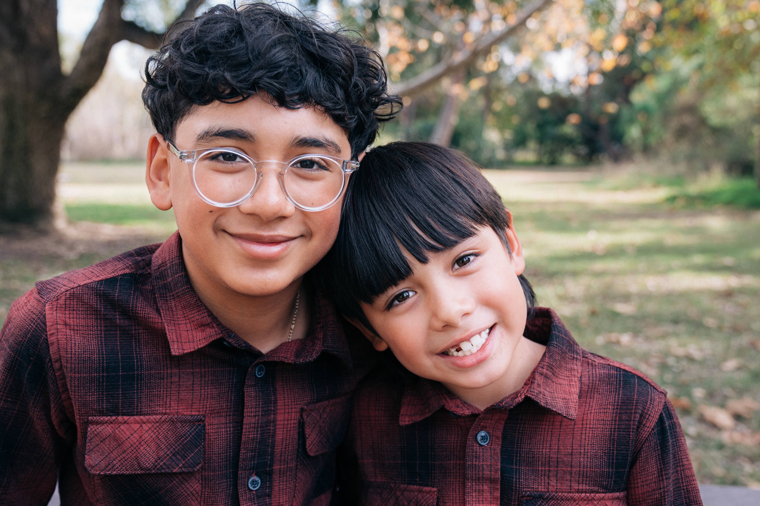 Two smiling boys wearing plaid shirts outdoors in a park with trees in the background.