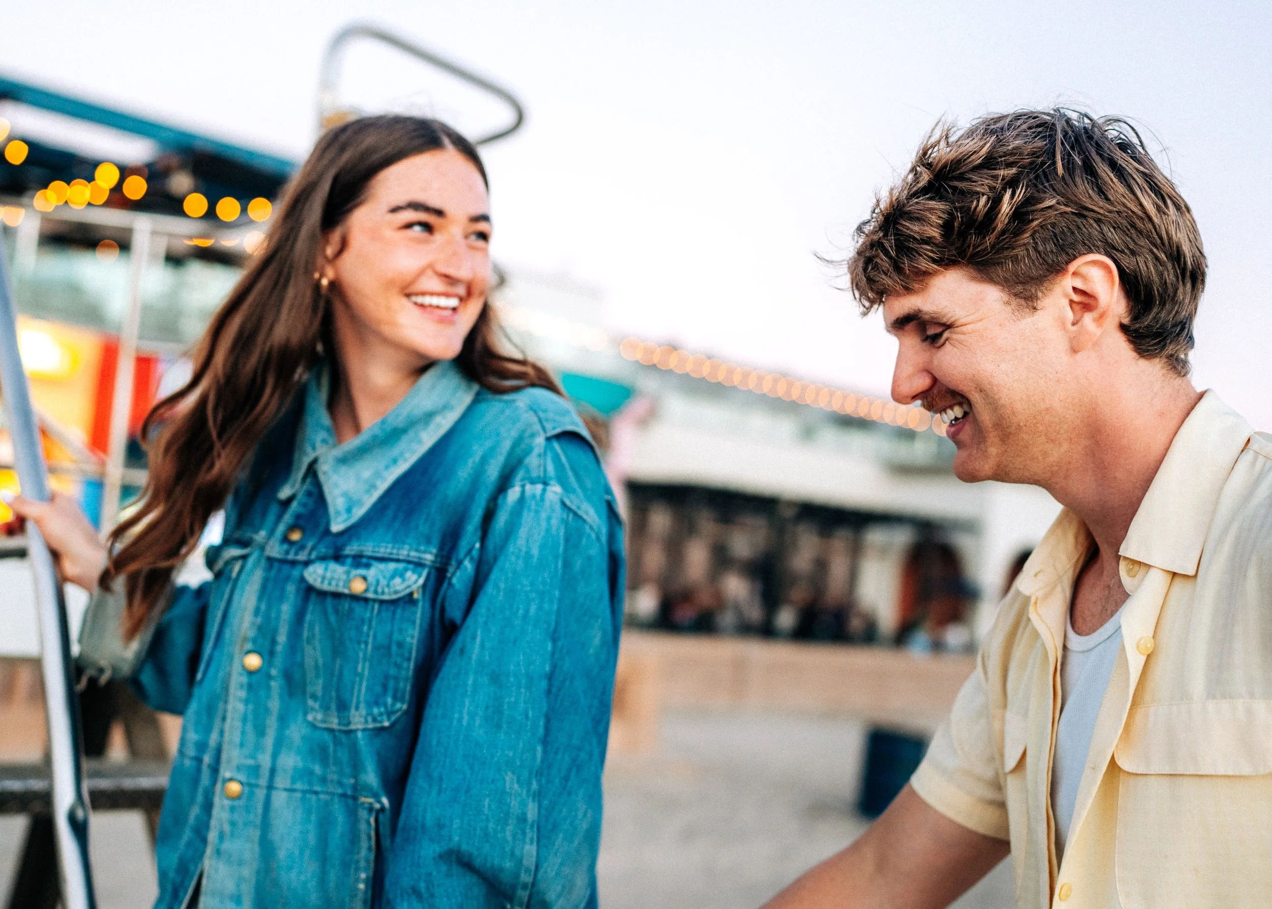 A young woman with long brown hair wearing a denim jacket smiling and talking to a young man with wavy brown hair wearing a light yellow shirt, at a fairground during daytime.