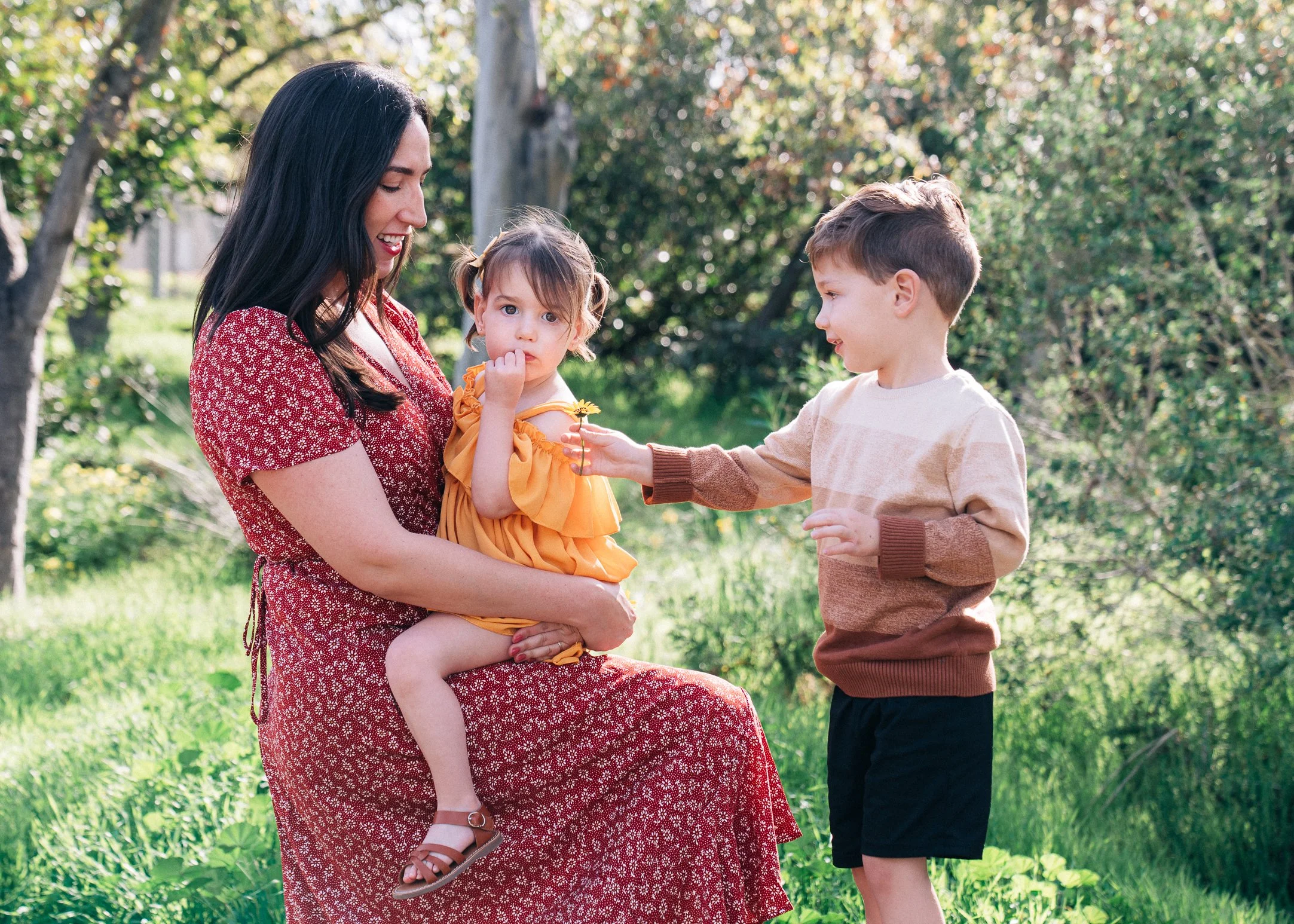 A woman holding a young girl sitting on her lap, with a boy standing in front of them in a park with green grass and trees.