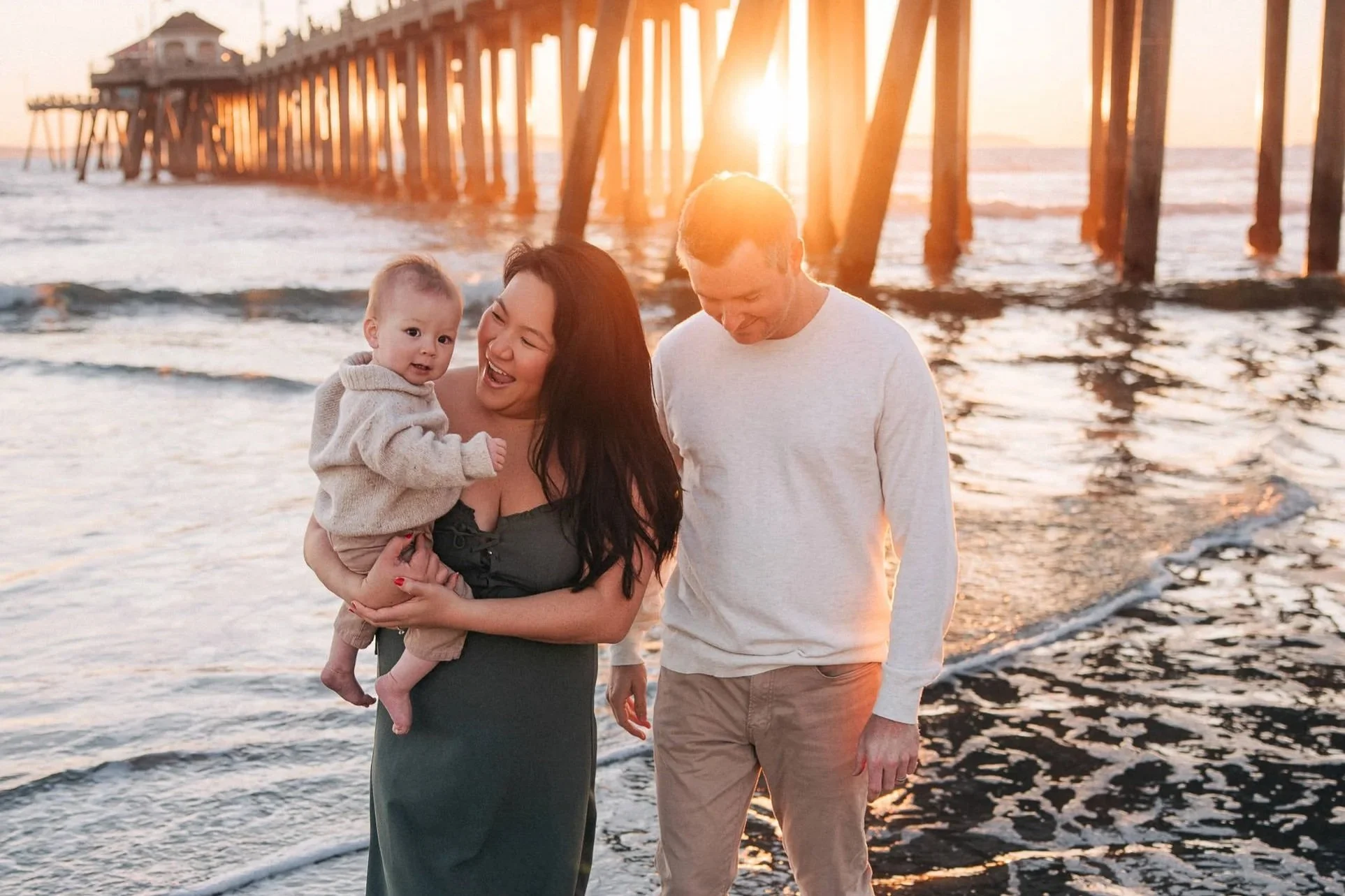 A family of three walking on the beach during sunset, with the mother holding a young child and the father walking beside them, the ocean and pier in the background.