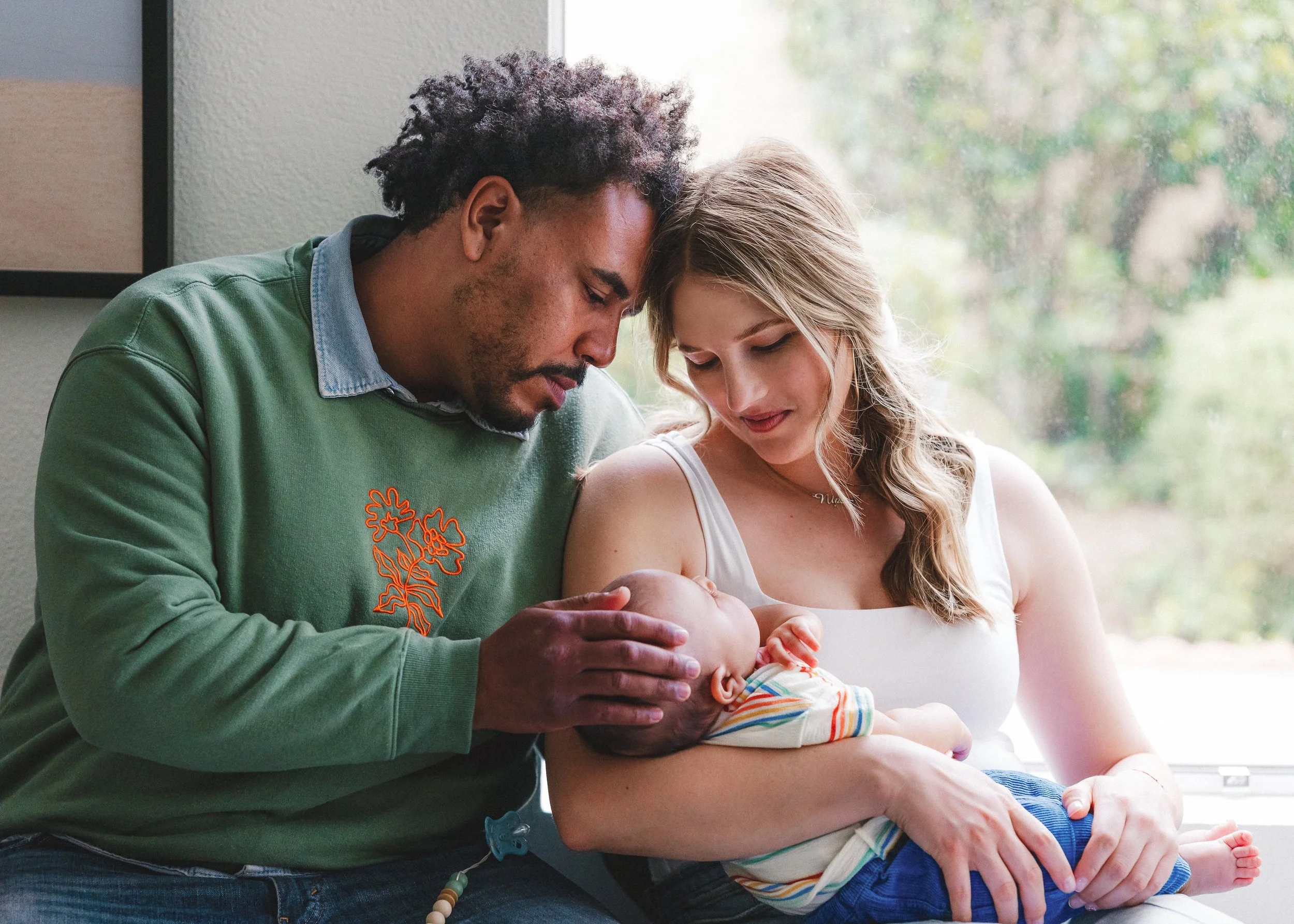 A diverse family in a hospital room: a man with curly hair, a woman with long blonde hair, and a newborn baby. The man gently touches the baby's head as the woman holds the baby on her lap.