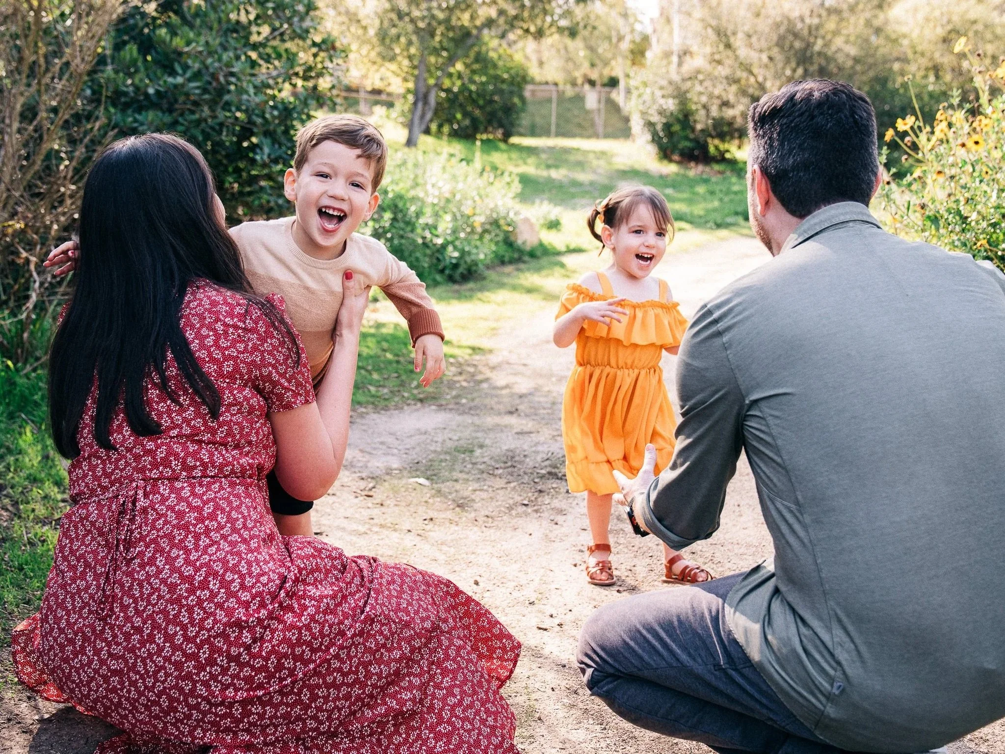 A family enjoying a moment outdoors on a sunny day, with a woman sitting on the ground wearing a red patterned dress, a young boy smiling and leaning on her, a little girl in an orange dress standing and laughing, and a man kneeling with his back to the camera, interacting with the girl, surrounded by greenery and trees.