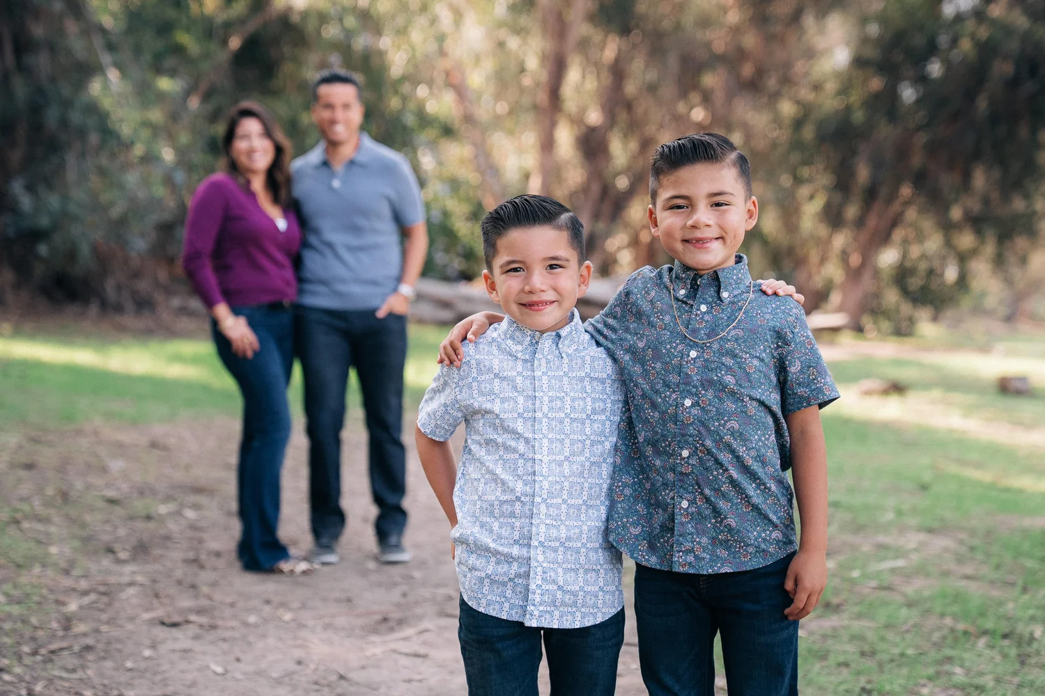 Two young brothers arm in arm on a shaded path at Gum Grove Park, their parents behind them