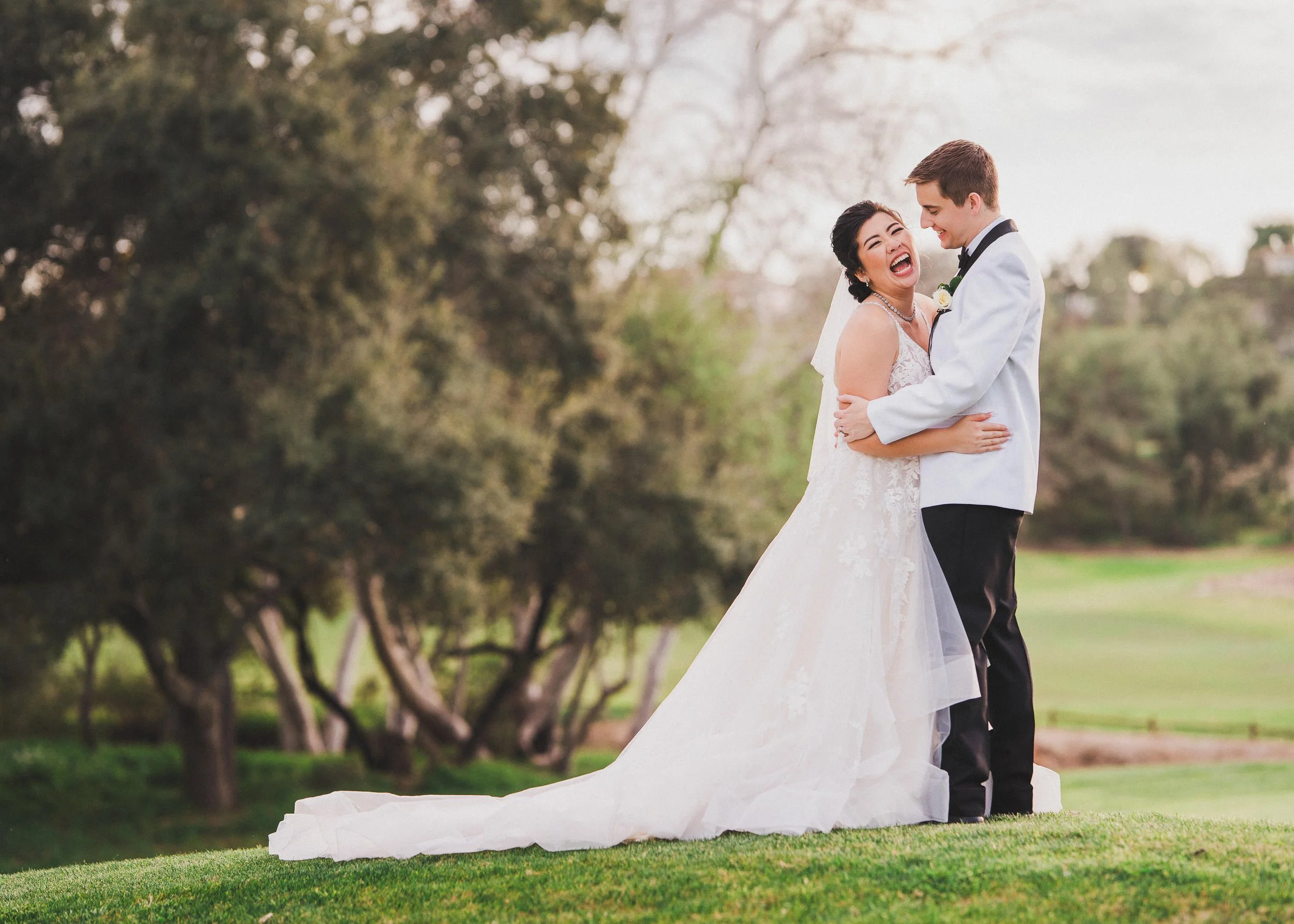 A bride and groom smiling and laughing in a park, with trees in the background, during their wedding photo shoot.