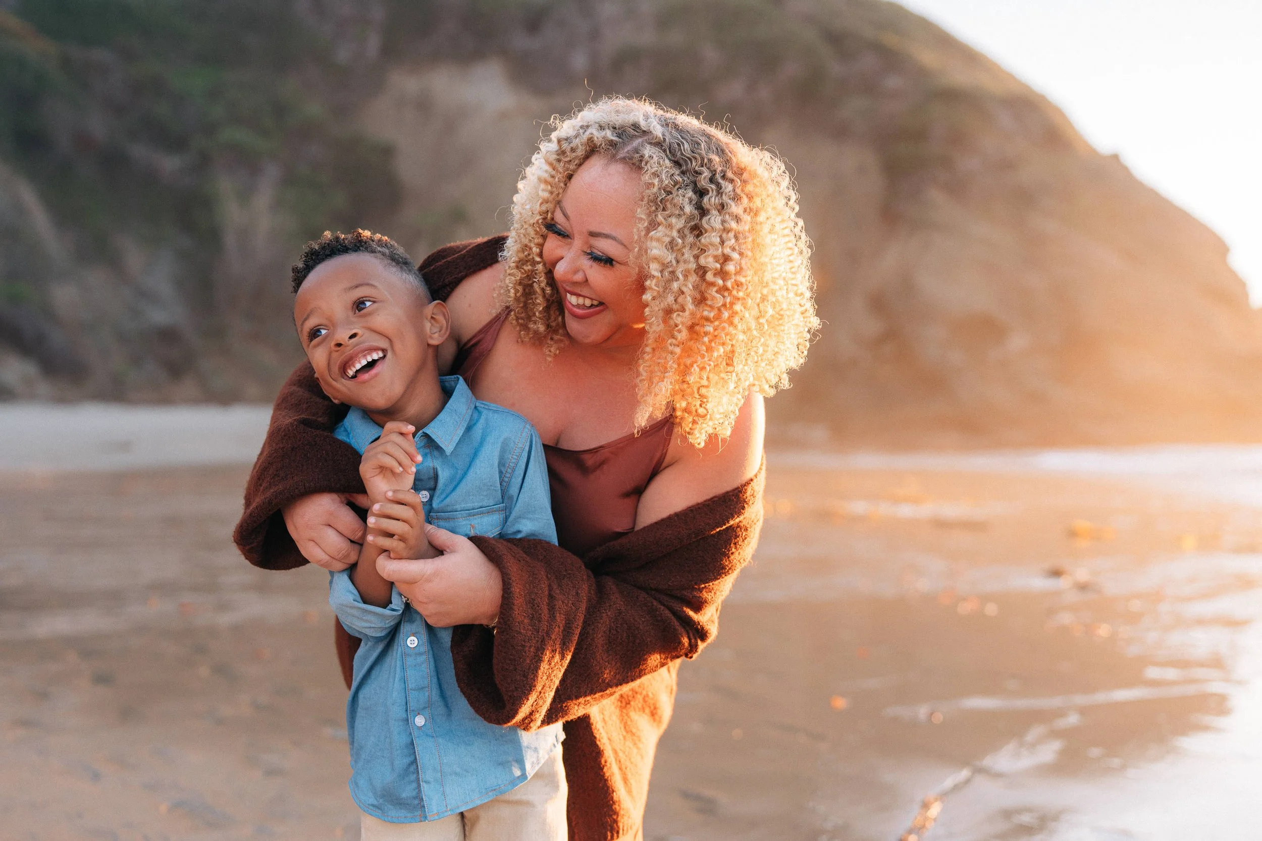 A woman and a young boy at the beach, smiling and laughing, with the woman hugging the boy. The background features a sandy beach and a large hill or cliff, with sunlight creating a warm glow.