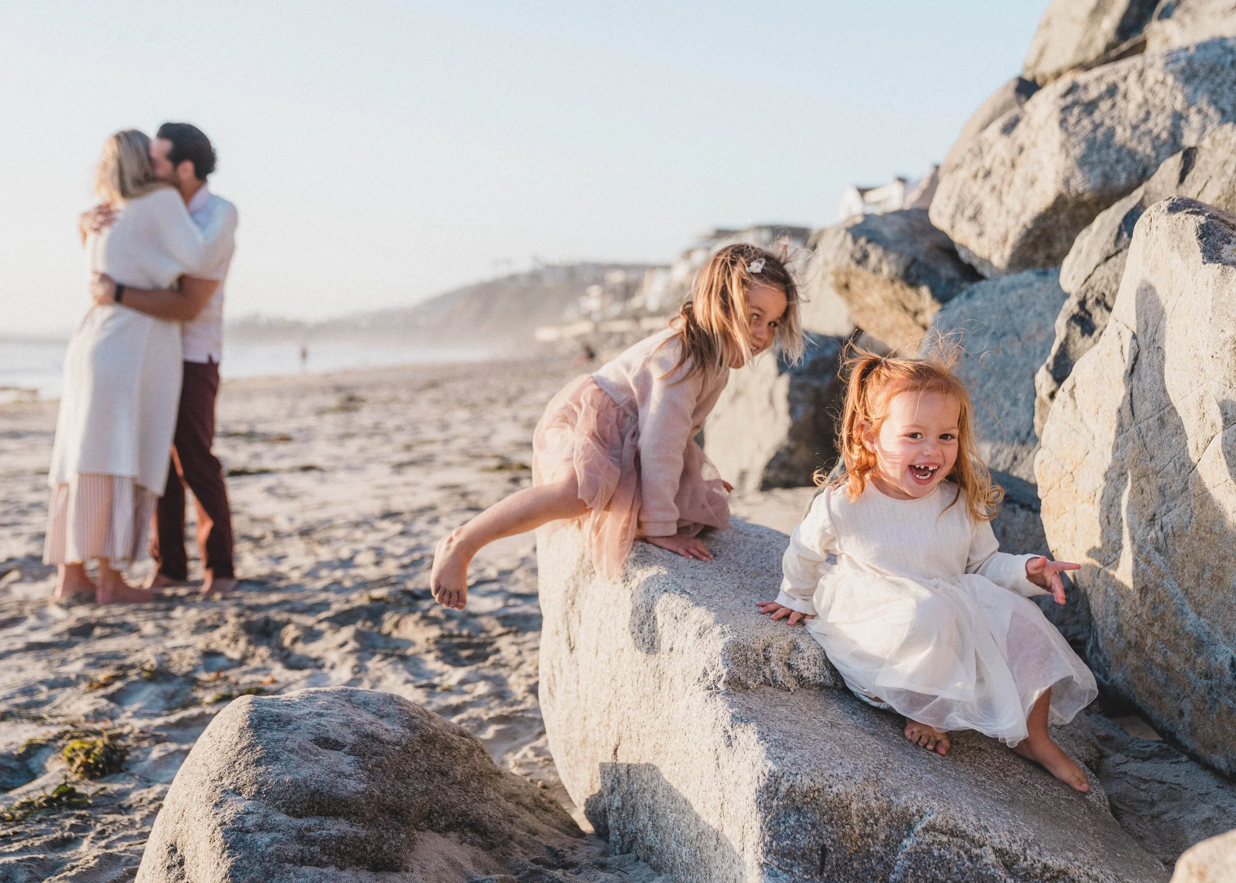 Two young girls with red hair playing on rocks at the beach with an older couple kissing in the background.