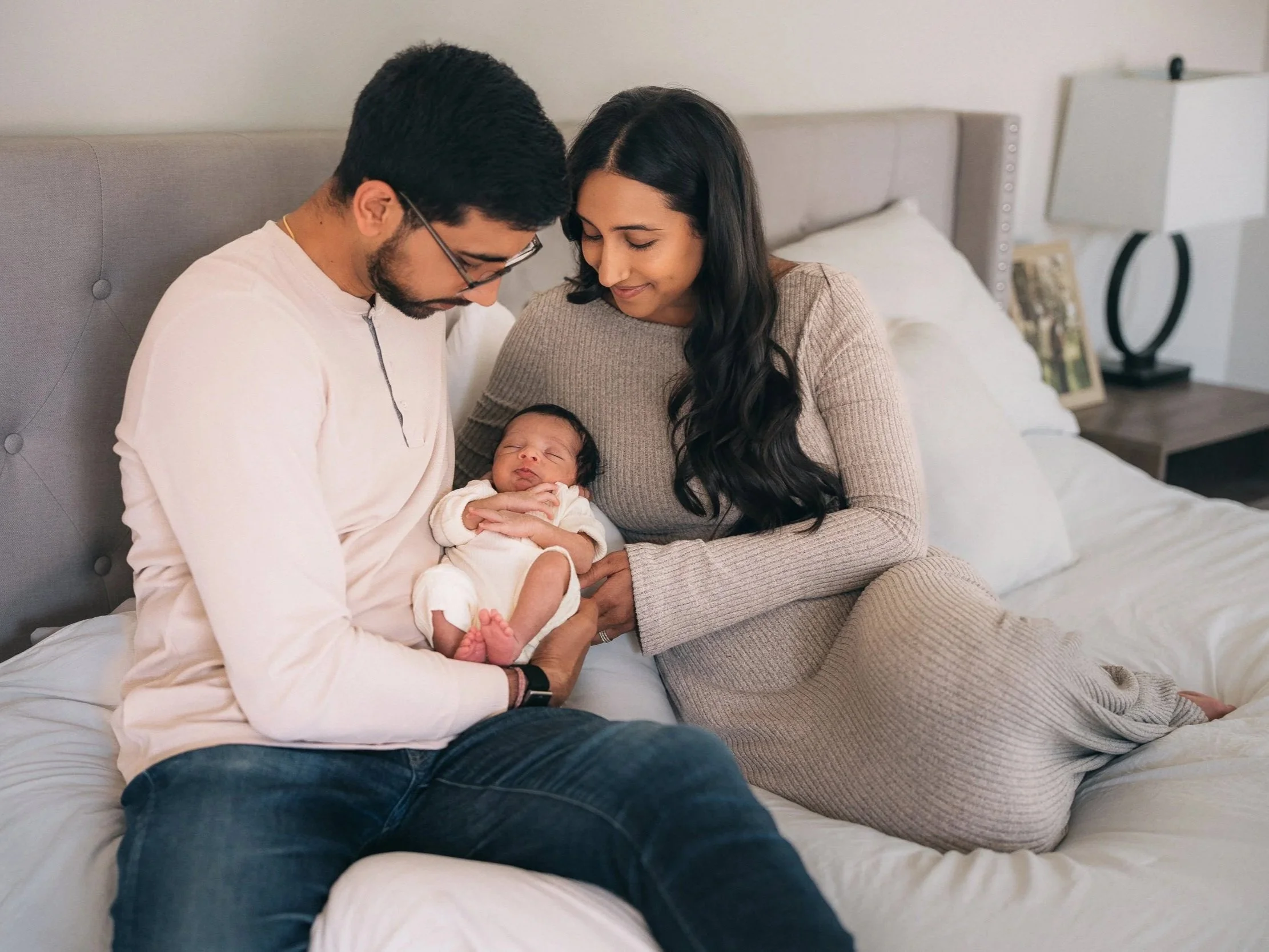A family of three sitting on a bed with a newborn baby. The father is wearing glasses and a white shirt, the mother has long dark hair and is wearing a beige sweater, and the baby is dressed in white, sleeping peacefully in the father's arms. They are in a cozy bedroom with a bedside table, lamp, and framed pictures in the background.