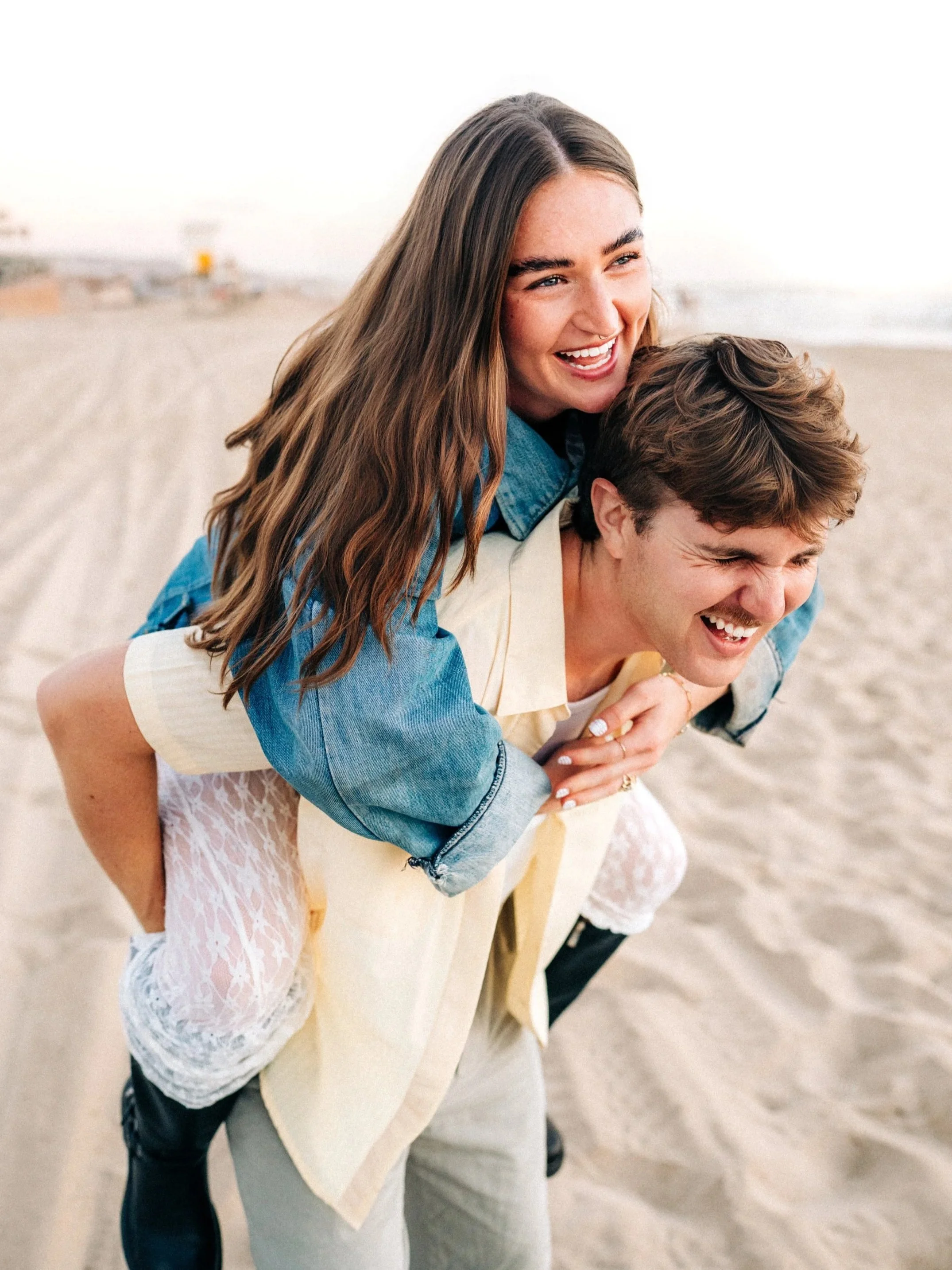 Two people on a beach, one giving the other a piggyback ride, both smiling and laughing.