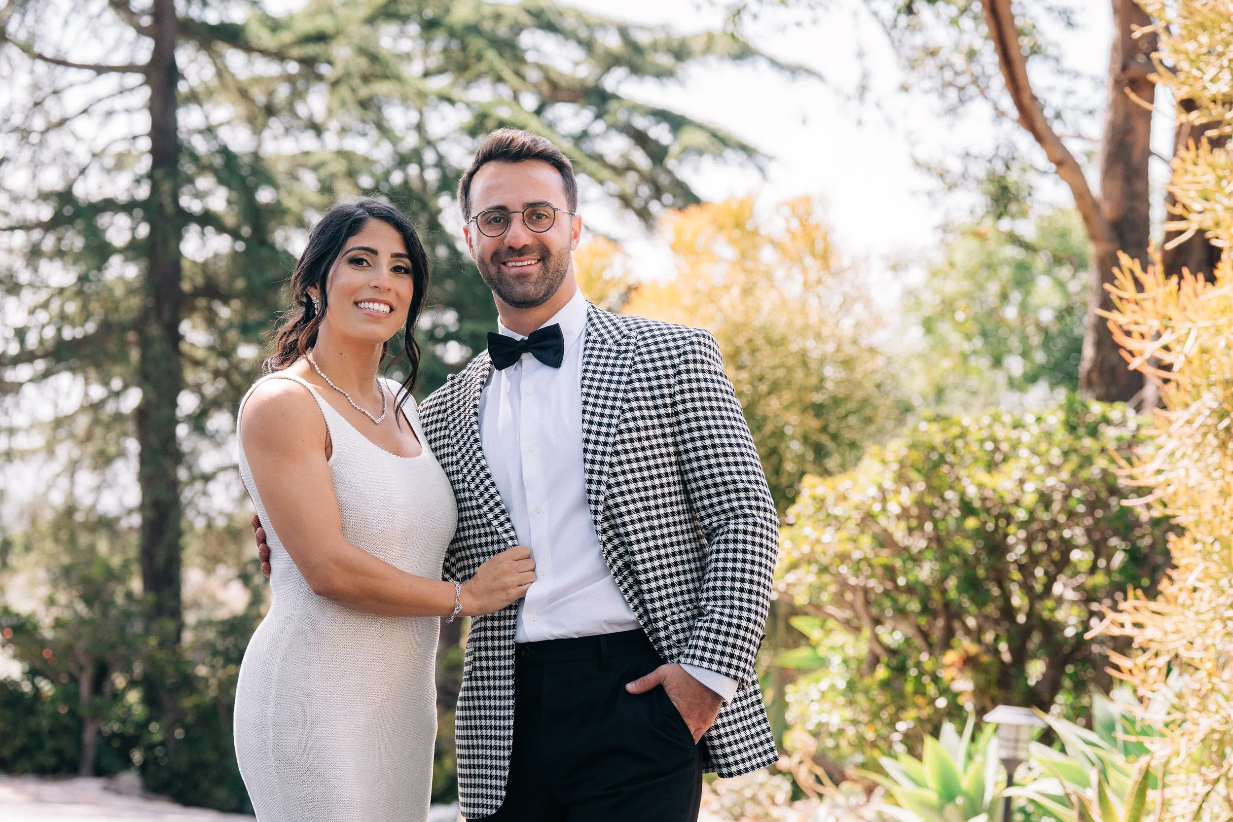 A smiling couple dressed up outdoors with trees and plants in the background, the woman in a white dress and pearl necklace, the man in a checkered blazer, white shirt, and bow tie.
