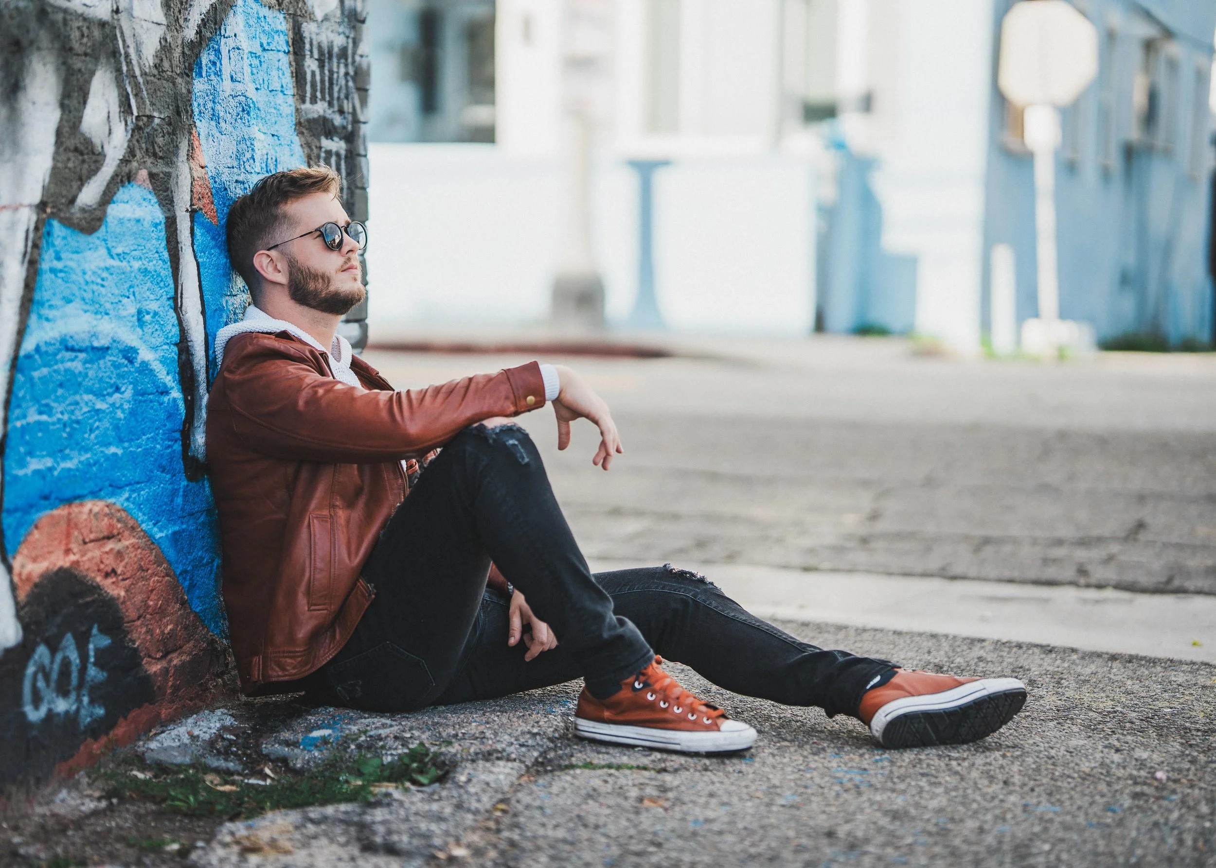 A young man with sunglasses, a brown leather jacket, black pants, and orange shoes sitting on the ground, leaning against a graffiti-covered wall in an urban alleyway.