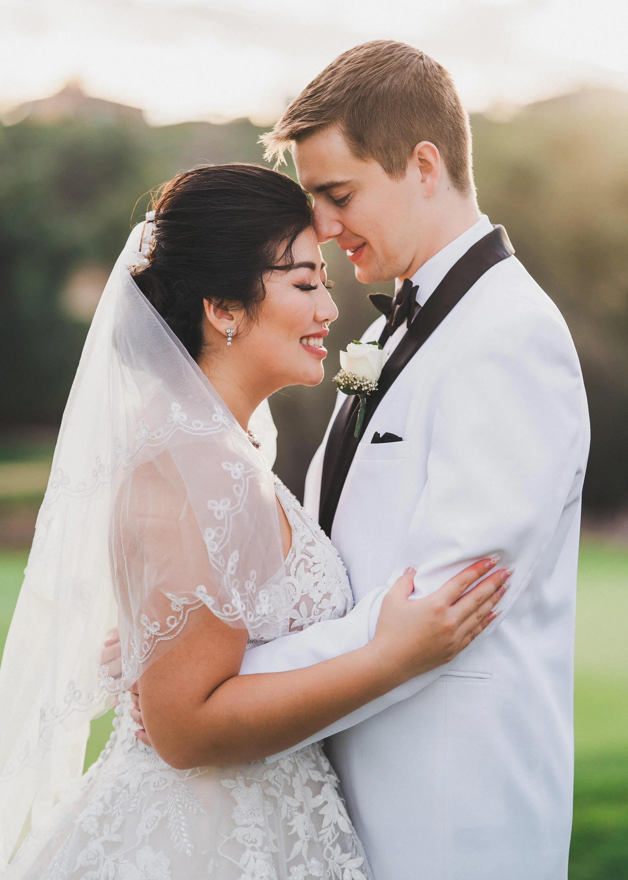 A bride and groom in wedding attire sharing a tender moment, with foreheads touching and eyes closed, outdoors during sunset.