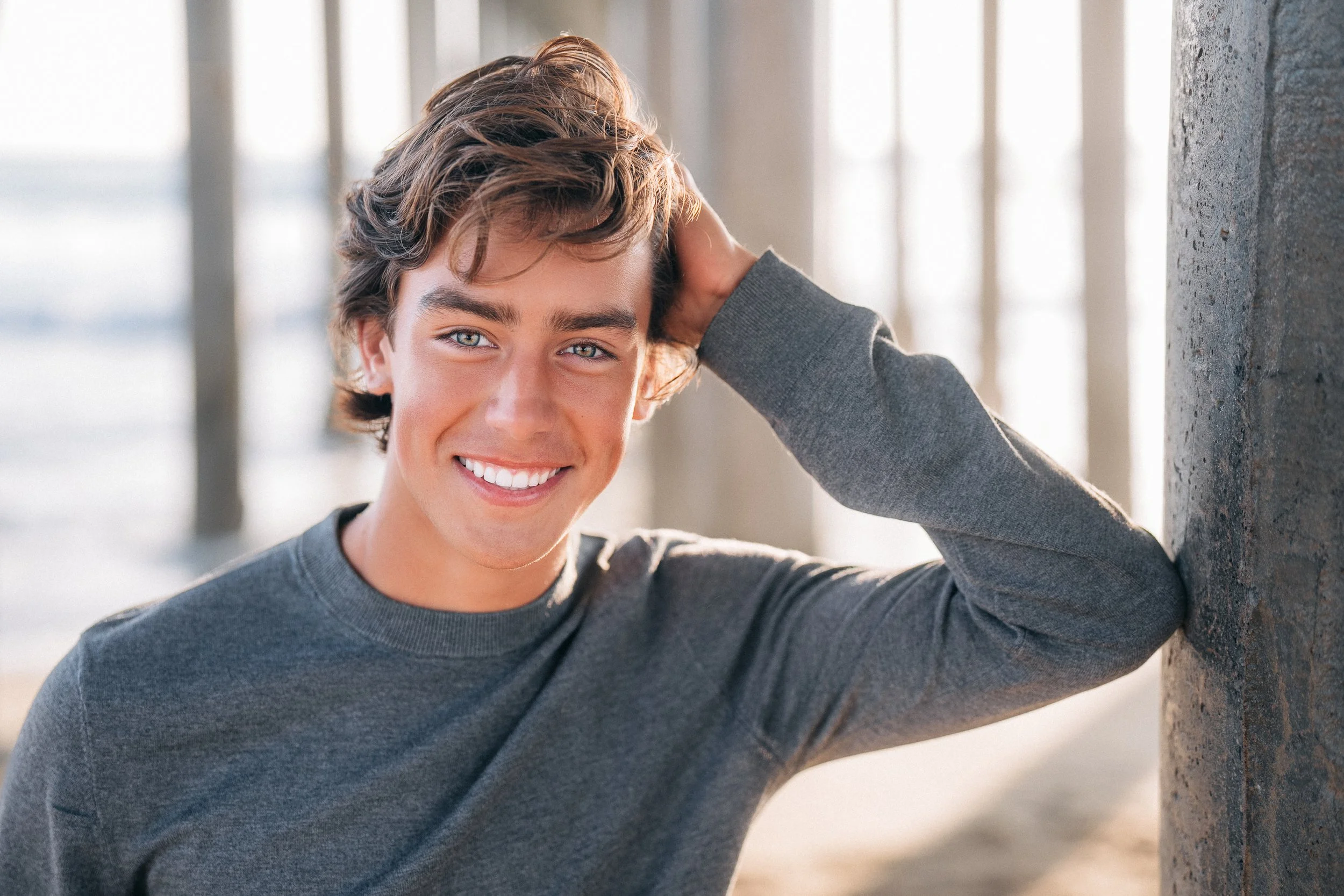 Smiling young man with wavy brown hair and blue eyes, wearing a gray long-sleeve shirt, leaning against a concrete wall with a blurry background of vertical columns.