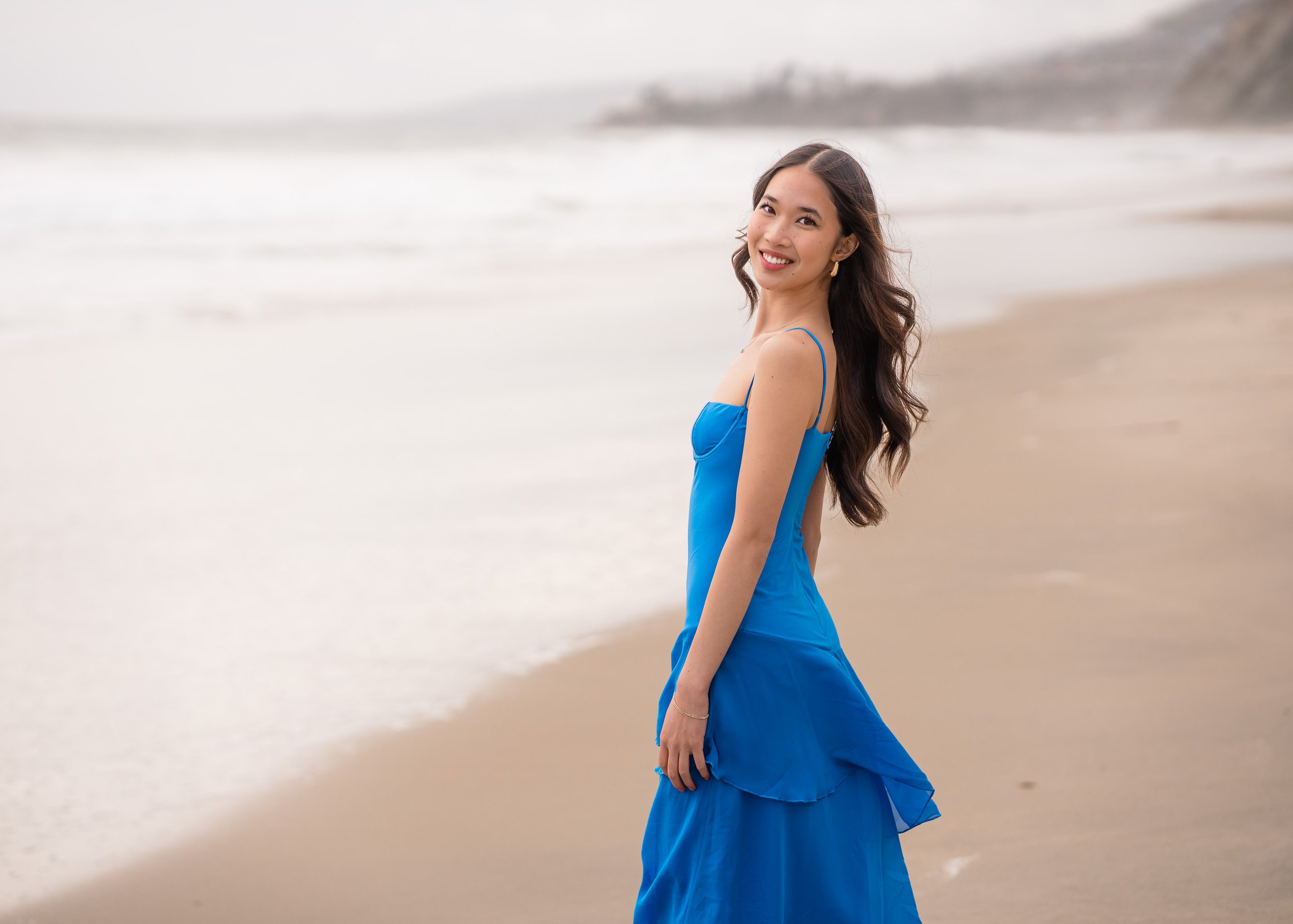 A woman in a long blue dress standing on a sandy beach with ocean waves and cliffs in the background, smiling at the camera.