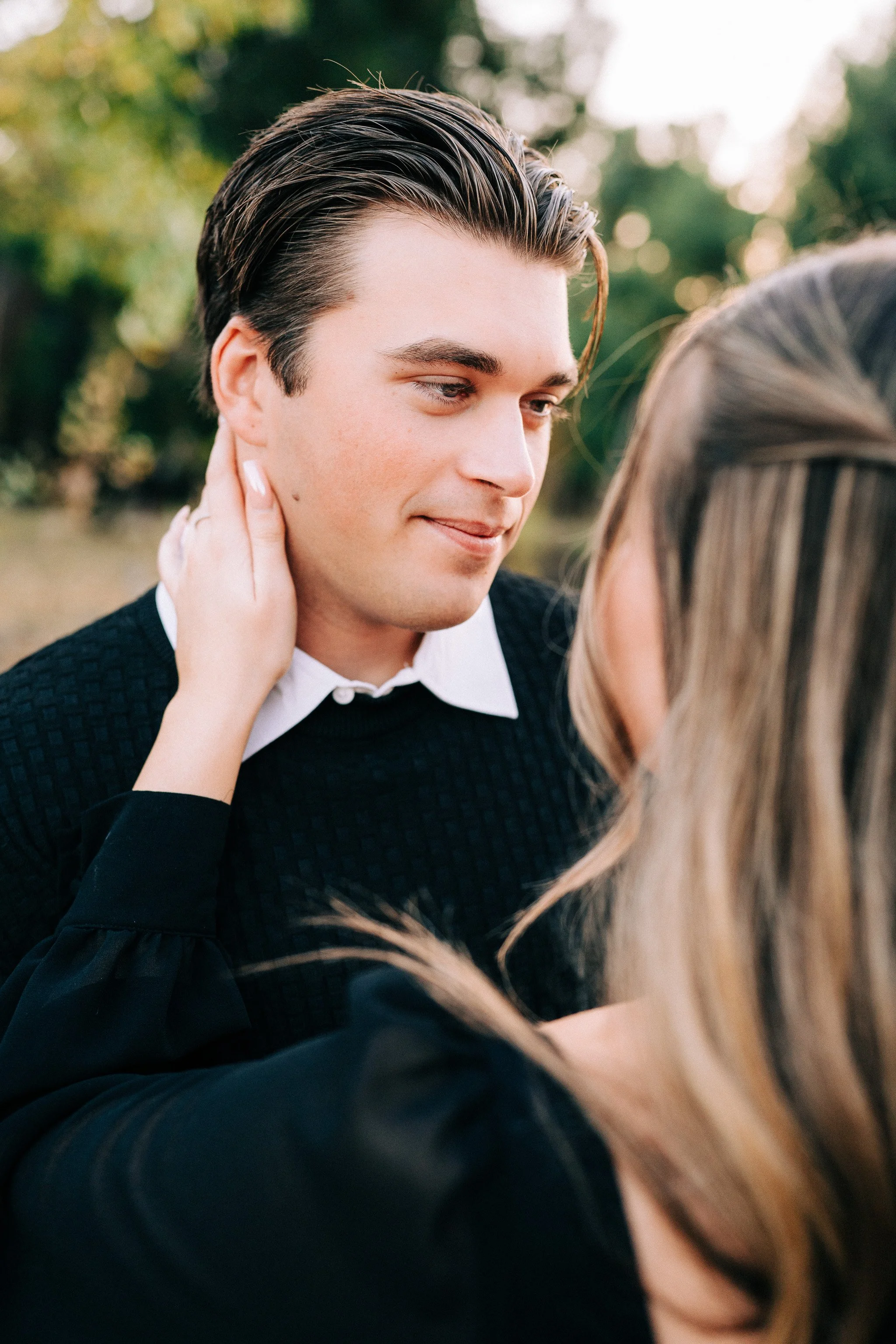 A young couple embraces outdoors, with the woman touching the man's neck and the man smiling softly, surrounded by blurred greenery.