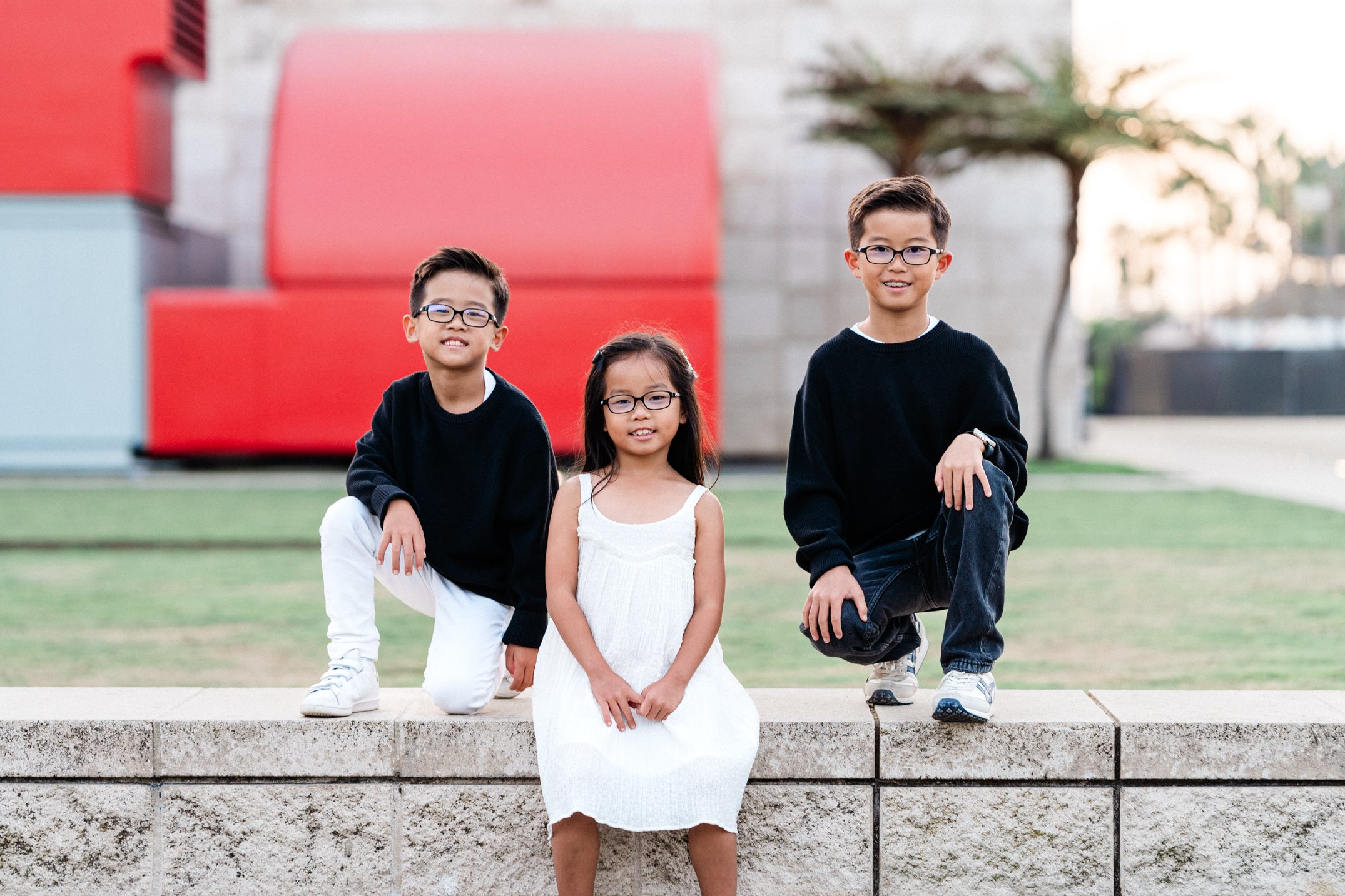 Four children, wearing glasses, are posing outdoors. The children are four, two boys and two girls. The girl in the front, in a white dress, is sitting on a concrete ledge, while the other three children stand behind her. The background includes a blurred sculpture and trees, with the sun in the distance.