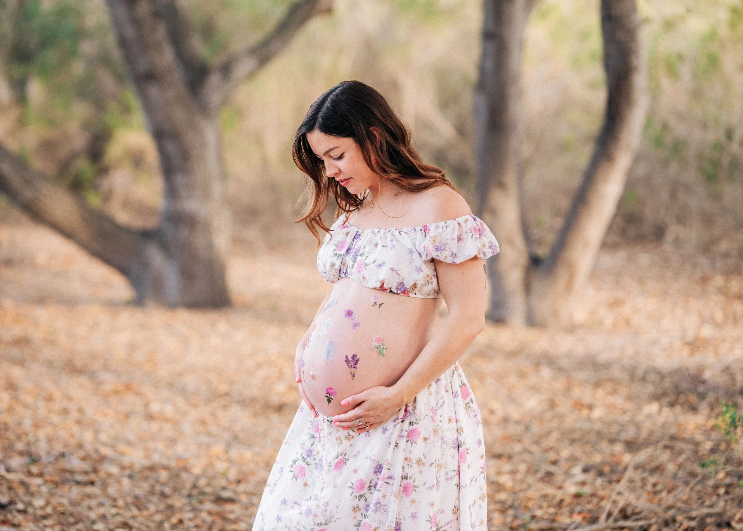 Pregnant woman in a floral dress with flowers painted on her belly, standing outdoors in a wooded area.